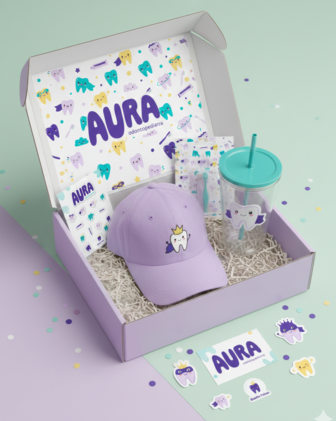 Caja de regalo con productos relacionados con la odontología infantil, incluye una gorra morada con una pequeña muela con corona, una taza con popote, stickers de dientes con diferentes expresiones y accesorios decorativos en tonos pasteles y con temática de dientes divertidos.