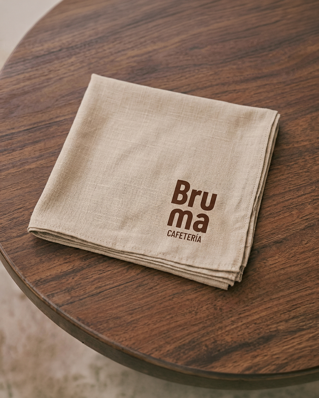 Servilleta beige doblada sobre mesa de madera con el logo 'Bru ma Cafetería' impreso.