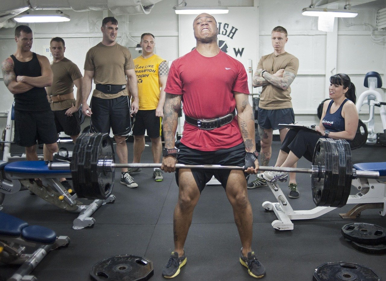 A man in a red shirt lifting a heavy barbell with weights in a gym, surrounded by six people watching him, some with arms crossed and one woman sitting on a workout bench smiling.