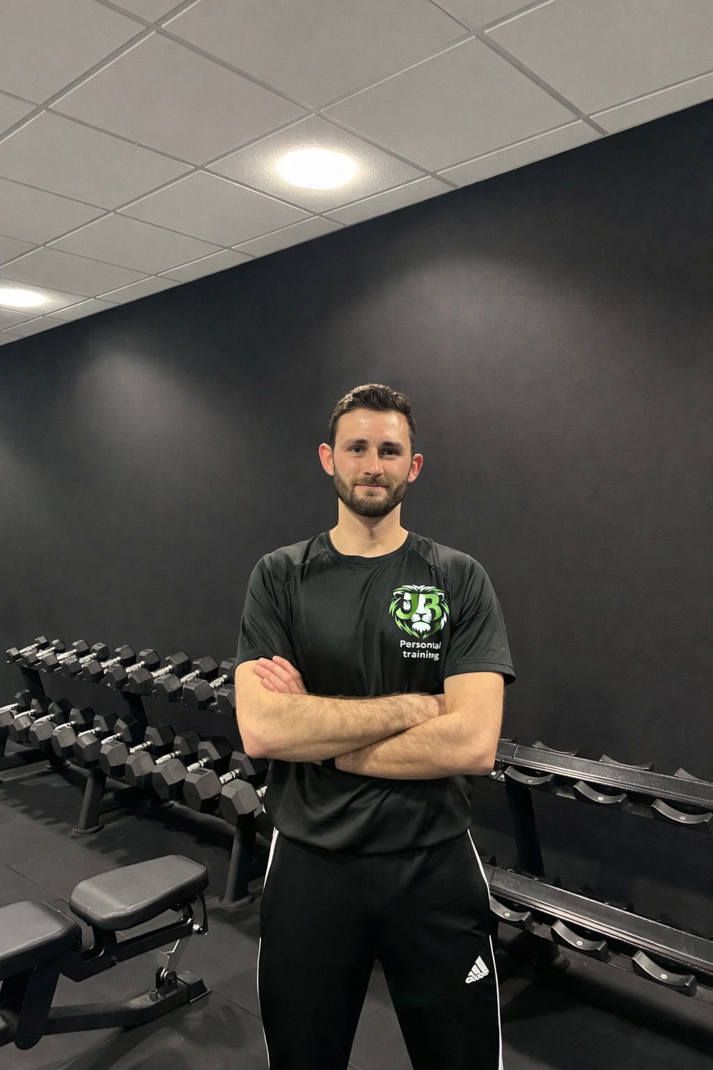 A man standing in a gym, wearing a black T-shirt and black pants with white stripes, crossing his arms, surrounded by dumbbells on racks.
