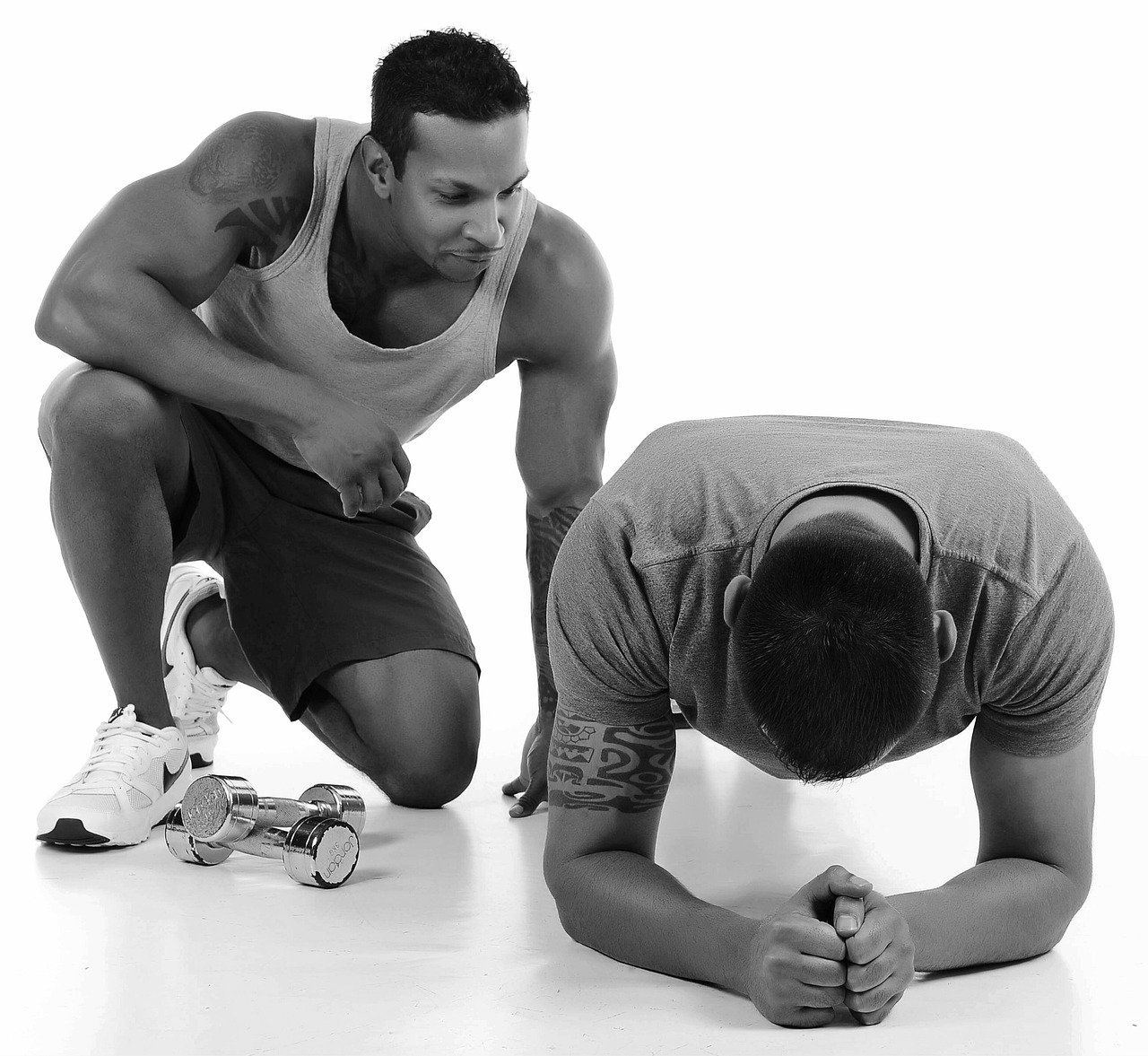 A man in sporty attire crouches nearby a second man who is doing an abdominal plank exercise on the floor, with dumbbells placed beside him.