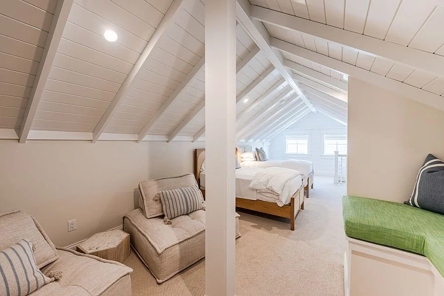 Bright attic bedroom with white paneled sloped ceilings, a bed with white bedding, a green cushioned built-in bench, and beige armchairs with striped pillows.