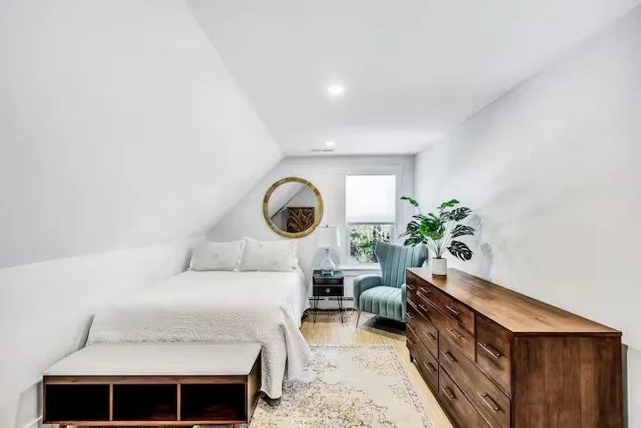 Cozy bedroom with white walls and sloped ceiling, king-sized bed with white bedding, round wall mirror, window with a view, blue armchair, wooden dresser, potted plant, bedside table with lamp, and area rug.