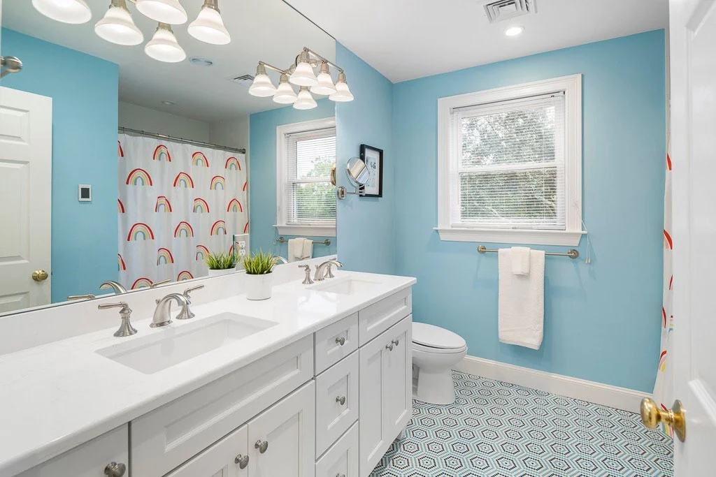 Bright bathroom with light blue walls, white vanity and double sinks, framed mirror, chrome fixtures, a window with blinds, a white toilet, and rainbow-themed shower curtain.
