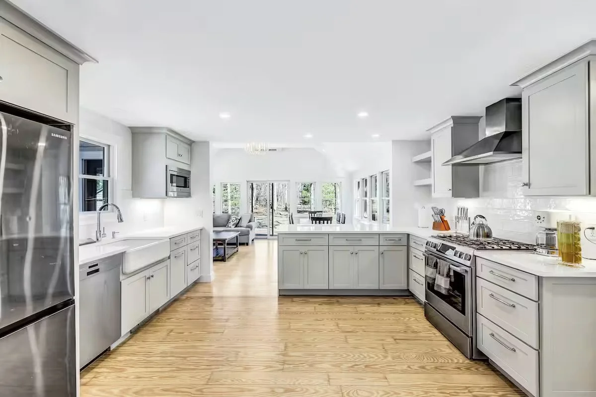 Bright, modern kitchen with white cabinets, stainless steel appliances, and hardwood floors, opening to a dining and living area with large windows and outdoor views.