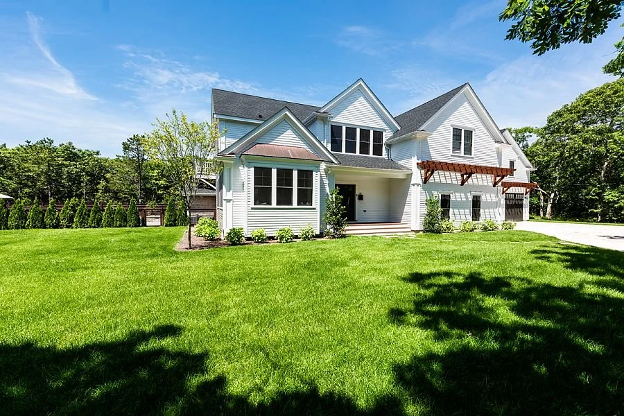 Large white house with multiple gabled roofs, surrounded by a lush green lawn and trees under a bright blue sky.
