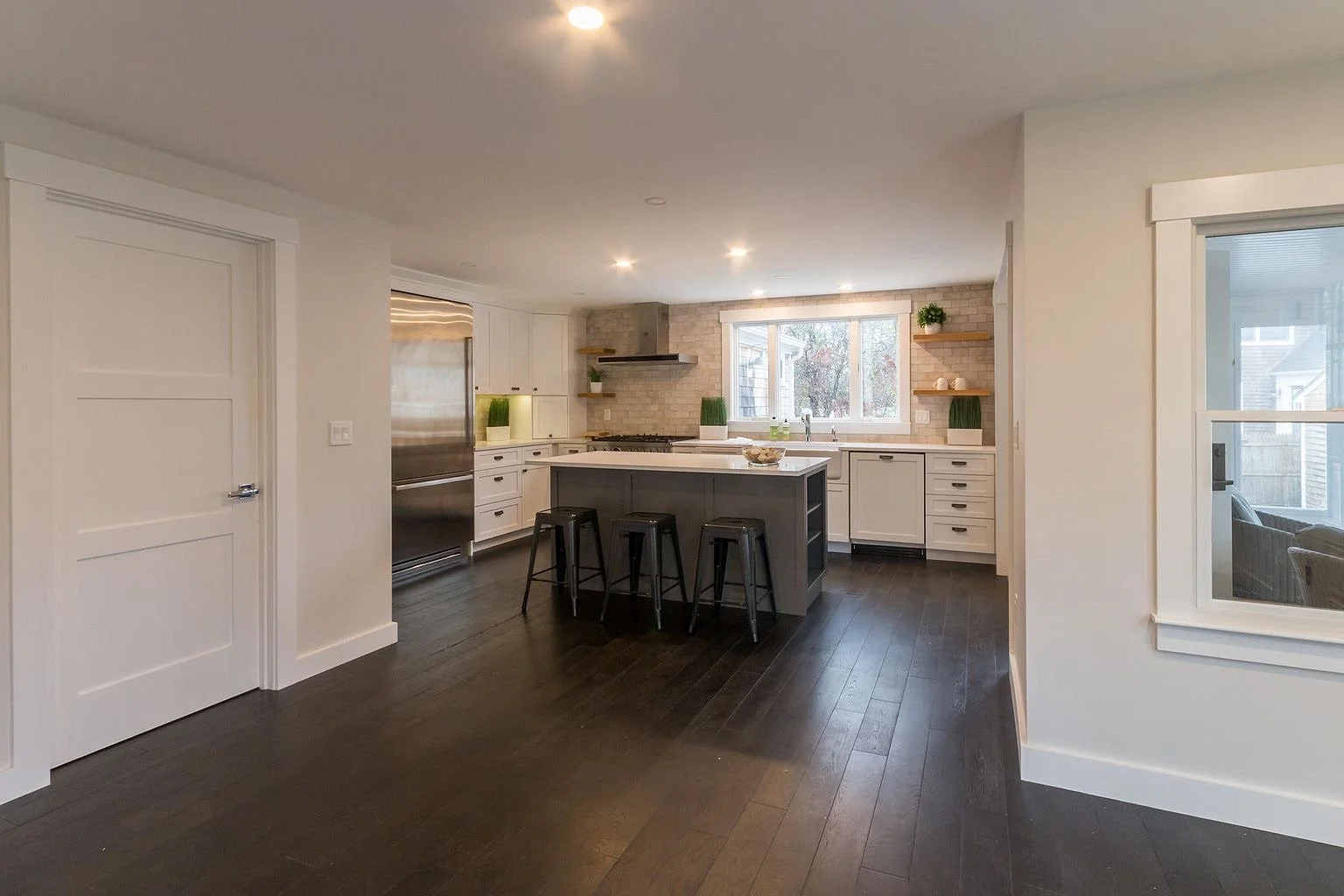 Modern kitchen with white cabinets, black countertop, kitchen island with three black stools, dark hardwood floors, large window, stainless steel refrigerator, brick backsplash, open shelving with green plants, and minimal decor.