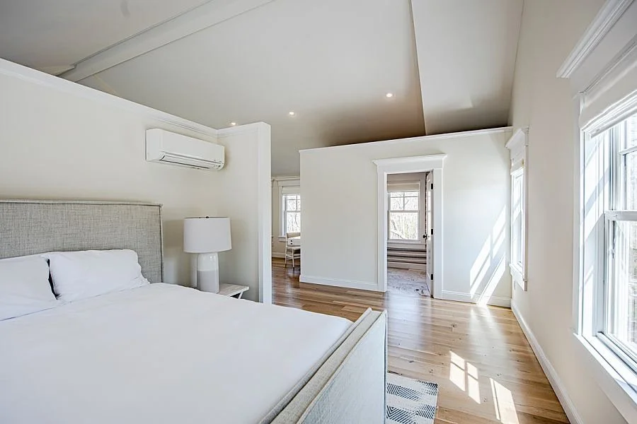 Bright, minimalist bedroom with a large bed, white linens, a nightstand with a lamp, hardwood floors, and windows letting in natural light.