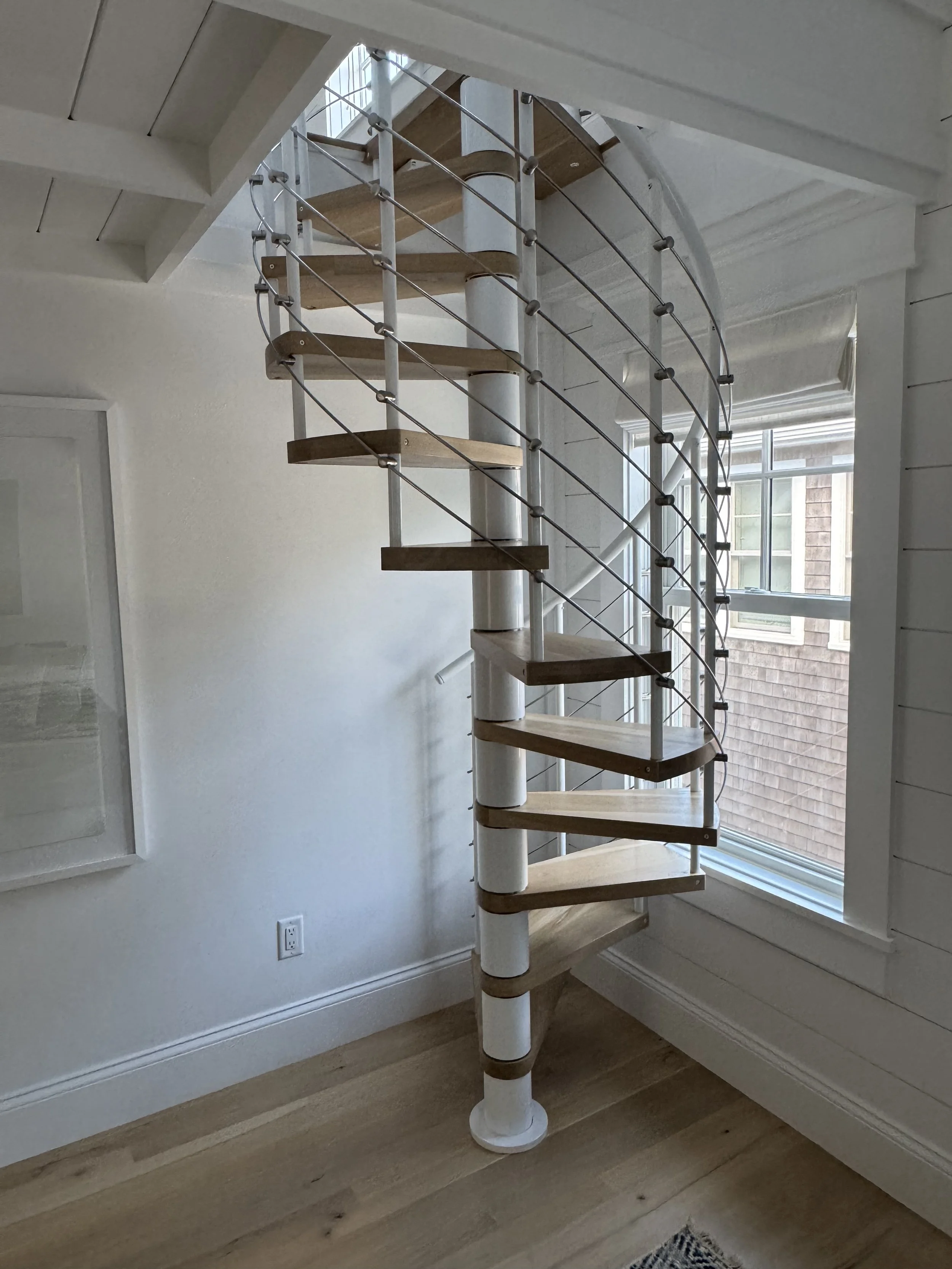 Indoor spiral staircase with wooden steps, metal railing, and white support column, next to a window.