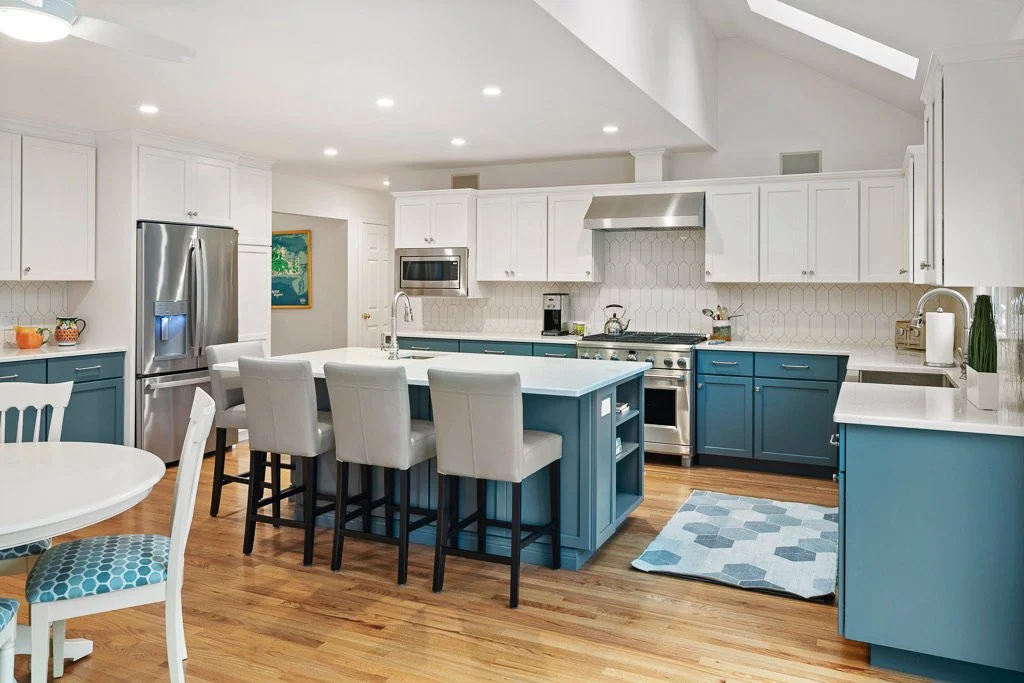 Modern kitchen with white upper cabinets, blue lower cabinets, stainless steel appliances, a kitchen island with barstools, and hardwood floors.