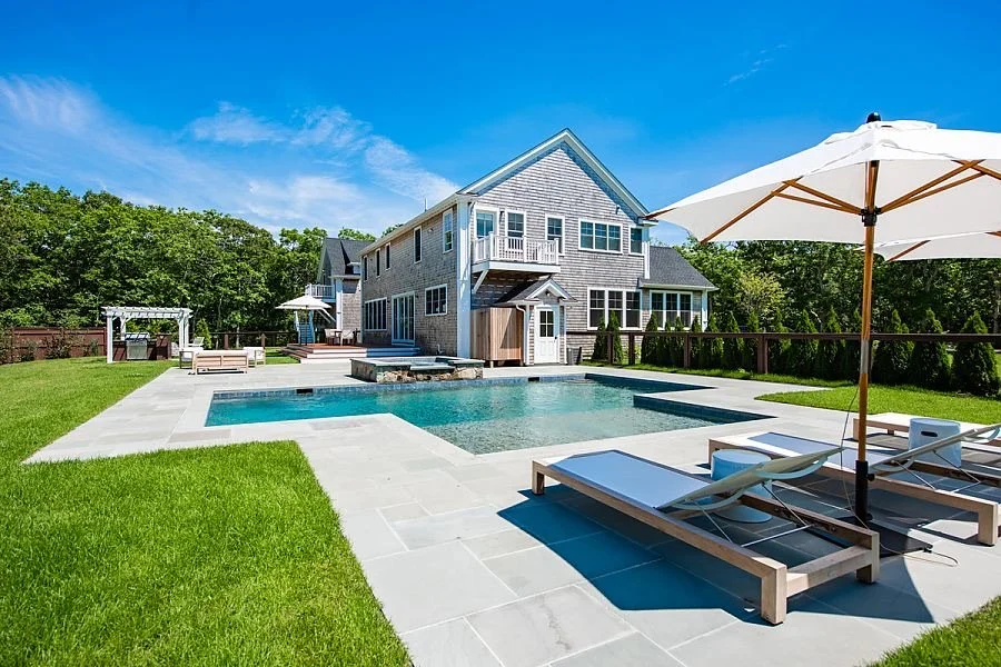 A backyard with a swimming pool, a well-maintained lawn, a house with gray siding, outdoor furniture, and a large white umbrella on a sunny day.