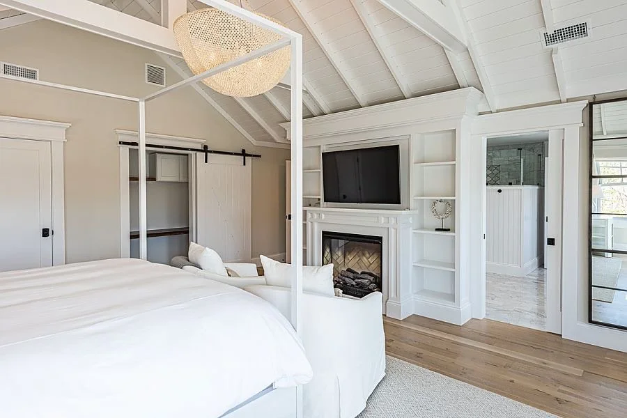Modern bedroom with white furniture, four-poster bed, built-in white shelves around fireplace, wall-mounted TV, and a sitting area with white cushions. The room features a vaulted ceiling with white wood paneling and a woven pendant light.