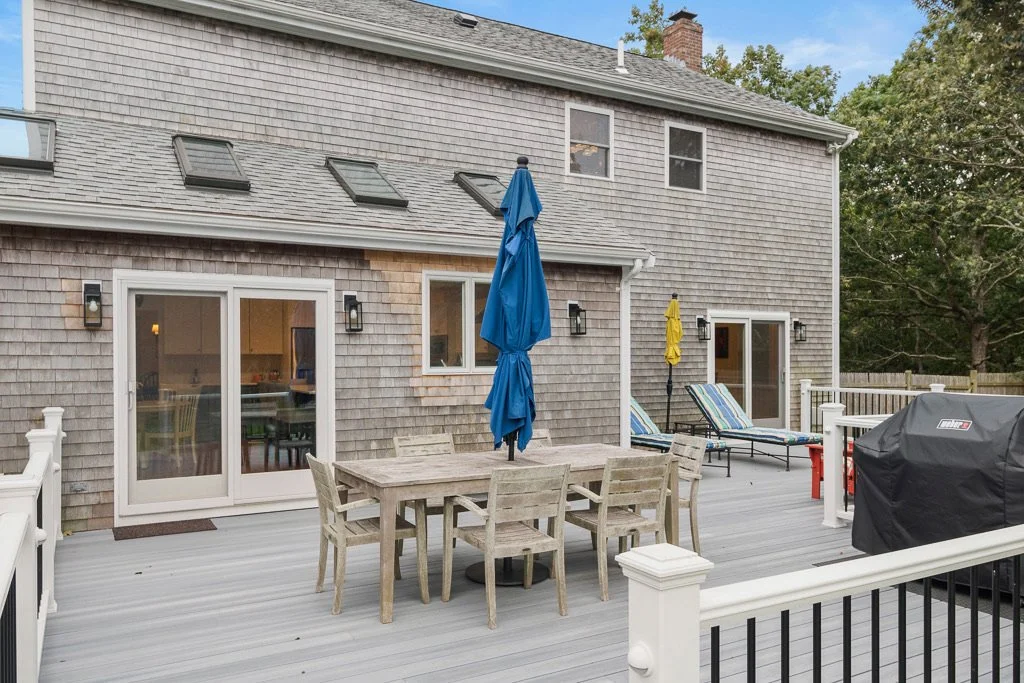 Outdoor deck with a wooden table, chairs, lounge chairs, umbrellas, a barbecue grill, and sliding glass doors leading inside a house.