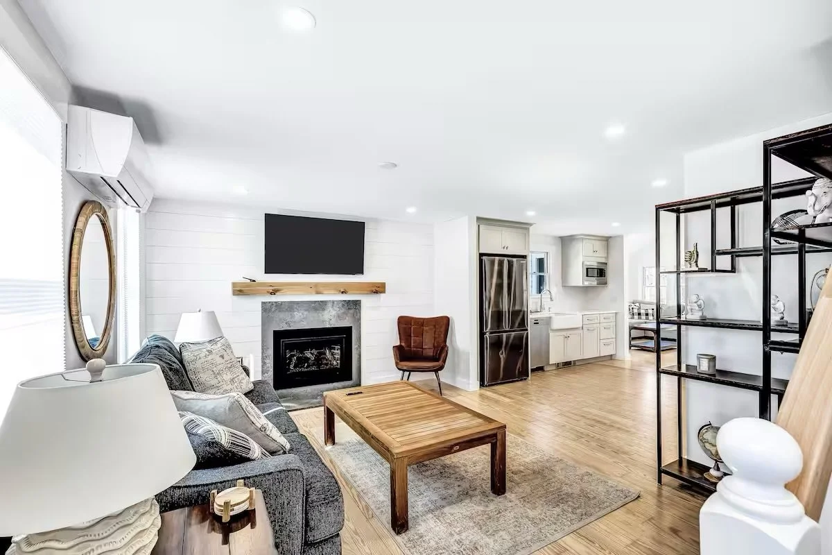 Open-concept living room with a sofa, wooden coffee table, armchair, a fireplace with a mounted flat-screen TV above it, and a metal shelving unit. The kitchen in the background features white cabinetry and stainless steel appliances.