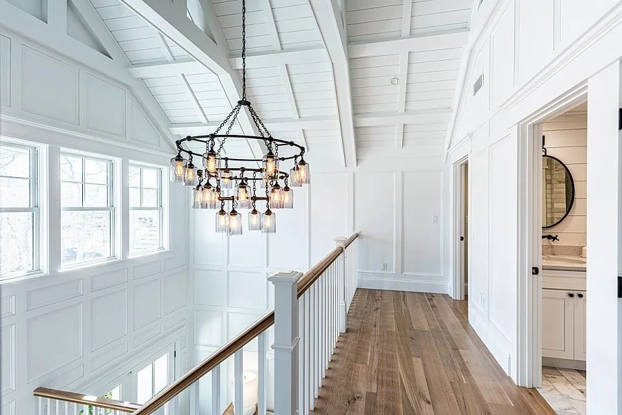 Bright upstairs hallway with white paneled walls, a wood railing, hardwood floors, and a black chandelier with multiple bulbs.
