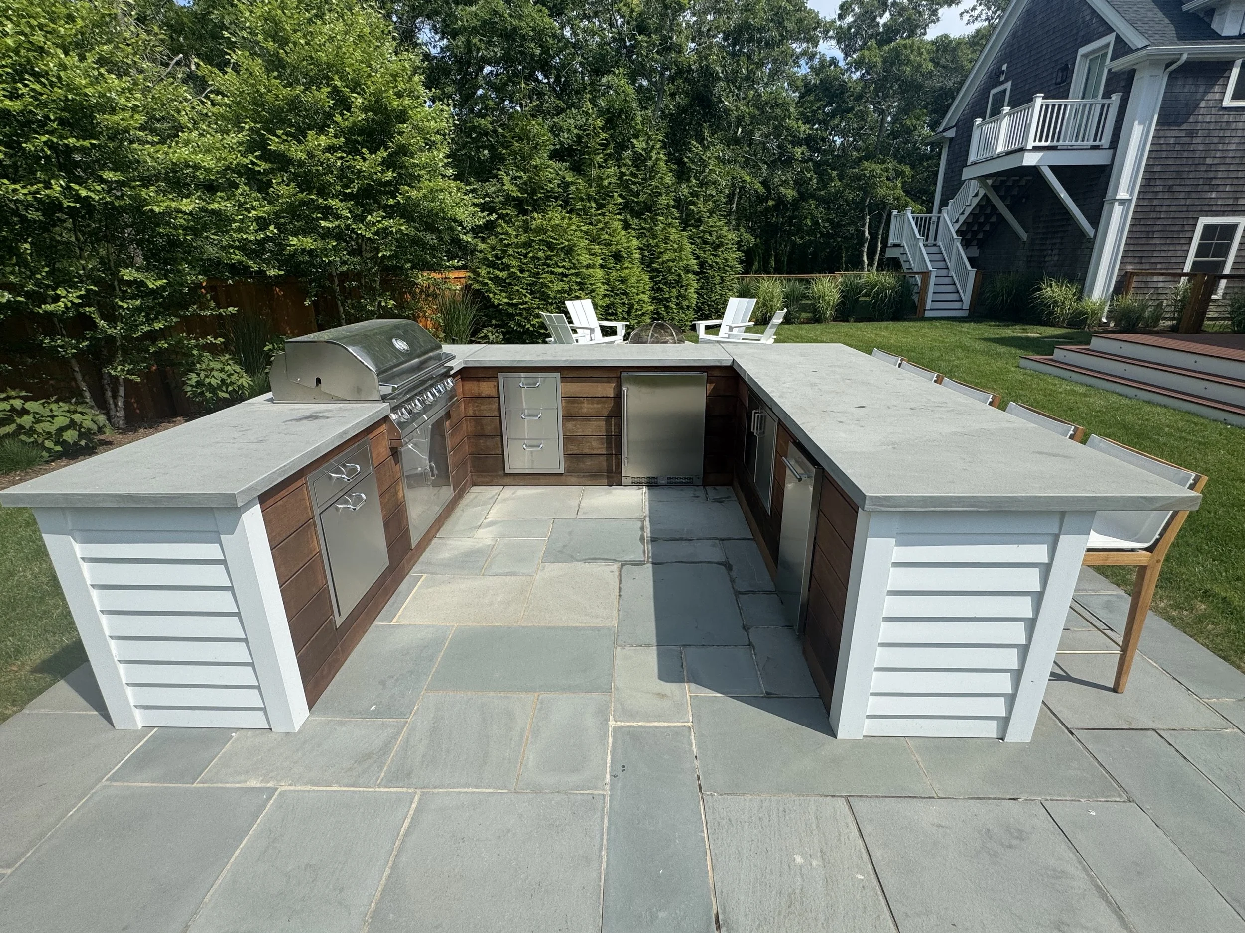 Outdoor kitchen with a large L-shaped counter, grill, and seating area, set on a stone patio in a backyard with trees, lawn, and a house in the background.