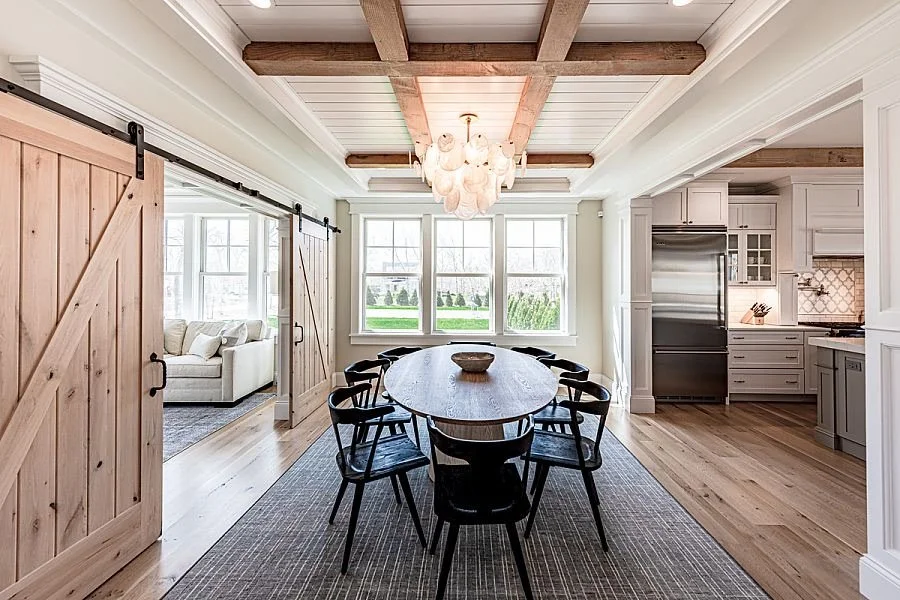 Dining room with large wooden table and black chairs, white kitchen with stainless steel appliances, barn door leading to living room, wooden beams and chandelier on ceiling, large windows with view of outdoors.