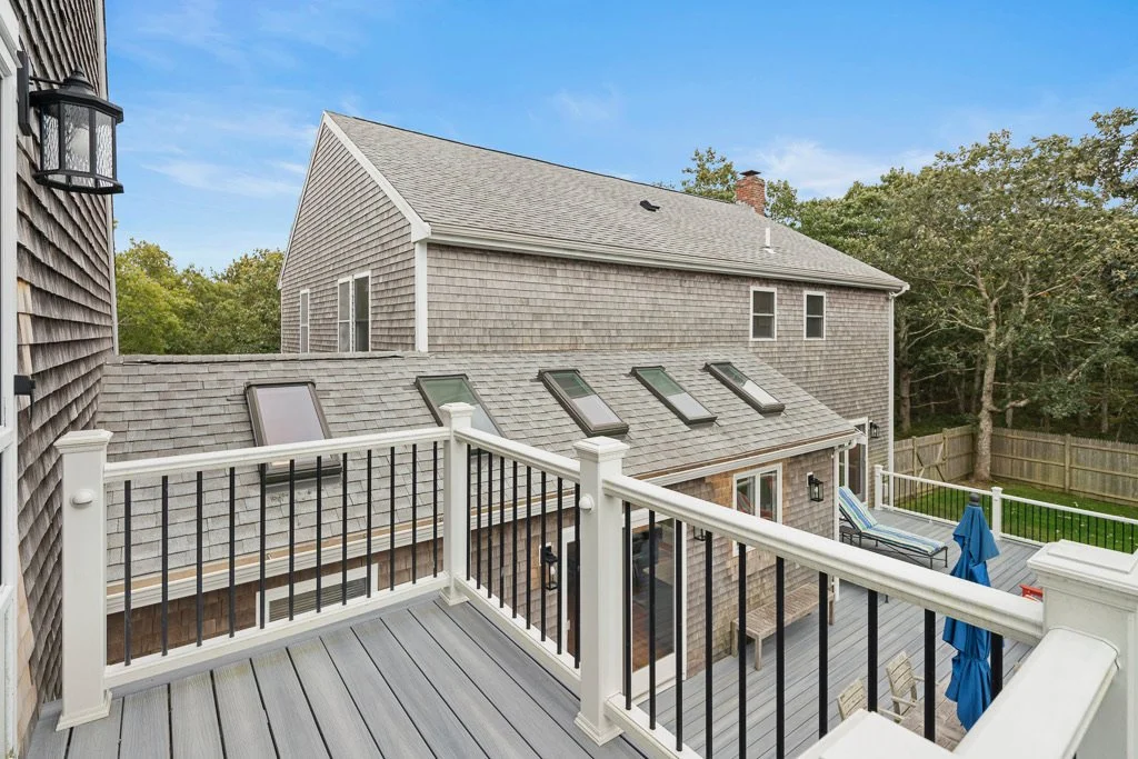 View of backyard deck with outdoor furniture and umbrellas, adjacent to a house with skylights, surrounded by trees and a wooden fence.