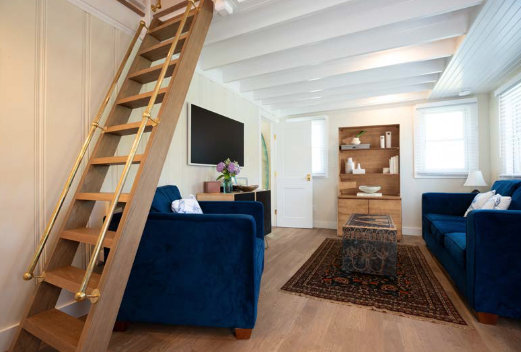 Living room with white ceiling beams, two navy sofas with white and blue pillows, wooden ladder leading to a loft, large windows with white blinds, a colorful rug, a wooden shelf with decorative items, a black TV, and a small side table with flowers.