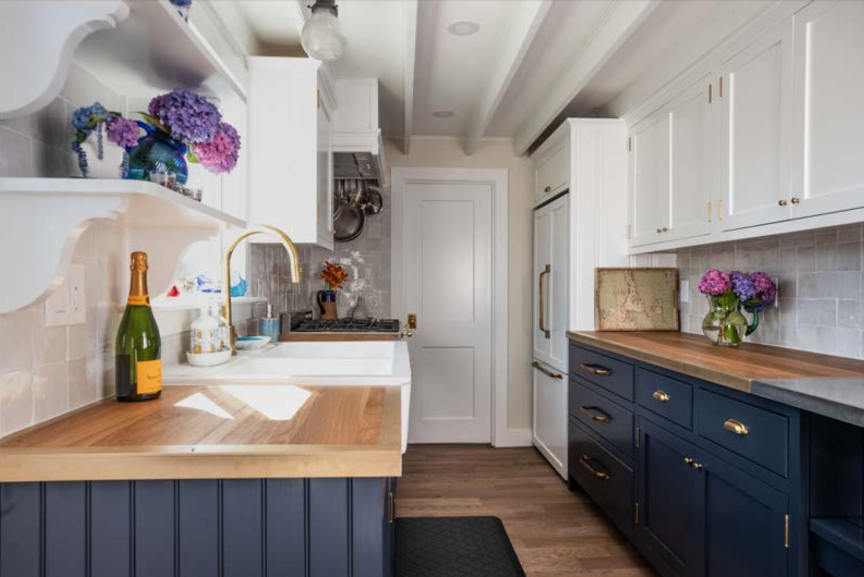 Kitchen with white upper cabinets, navy lower cabinets, wooden countertops, flower vases with pink and purple flowers, a bottle of champagne, and a window over the sink.