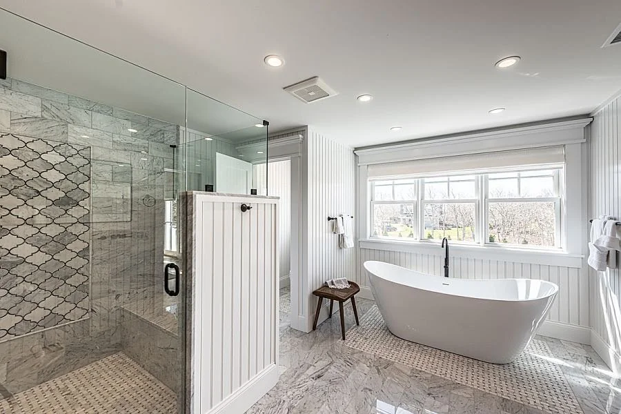 Bright bathroom with a freestanding bathtub, large window with white blinds, and a glass-enclosed shower with patterned tiles.