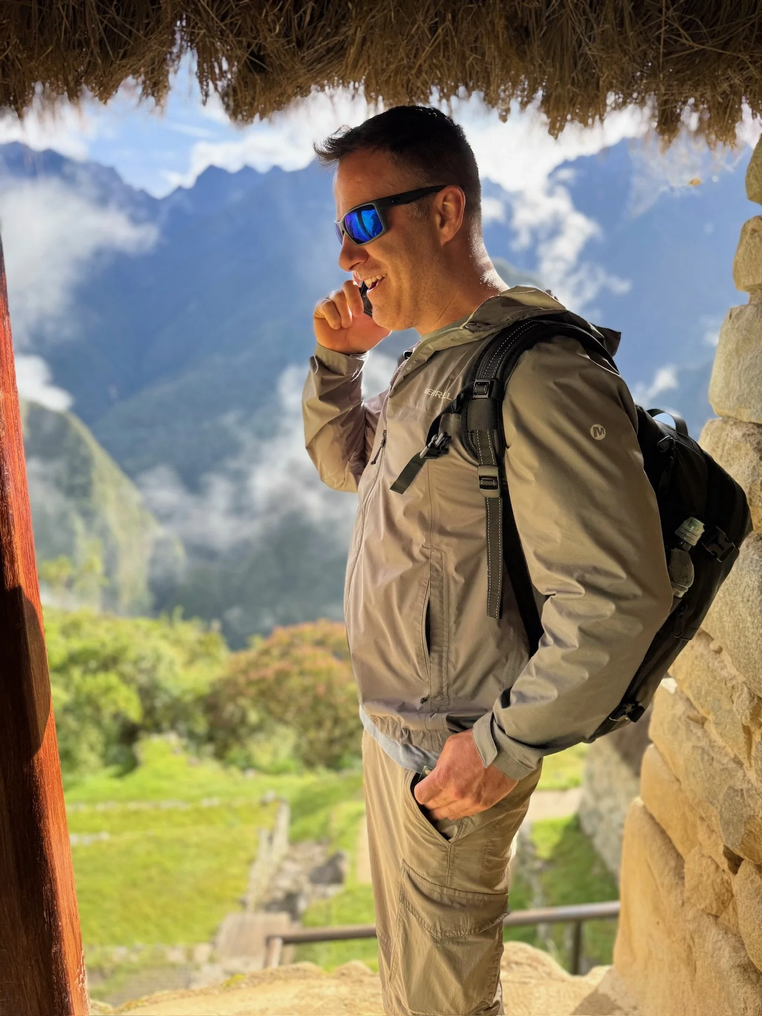 A man in outdoor gear, wearing sunglasses and a backpack, is talking on a cellphone while standing under a thatched roof with mountains and lush greenery in the background.