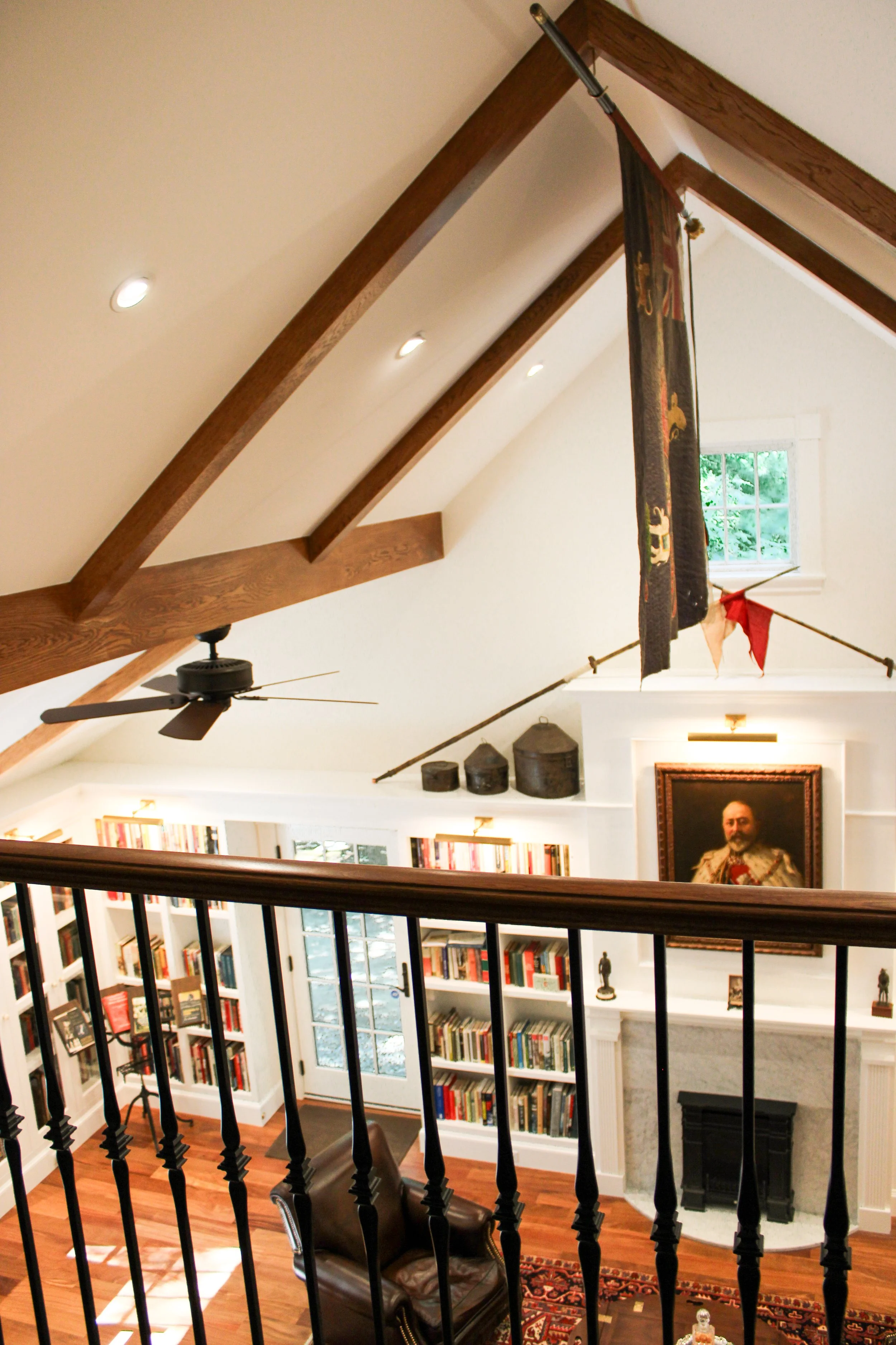 A view of a cozy living room from an upper floor, featuring bookshelves filled with books, a fireplace, a portrait painting on the wall, and a small window allowing natural light, with wooden beams, a ceiling fan, and a balcony railing in the foregro