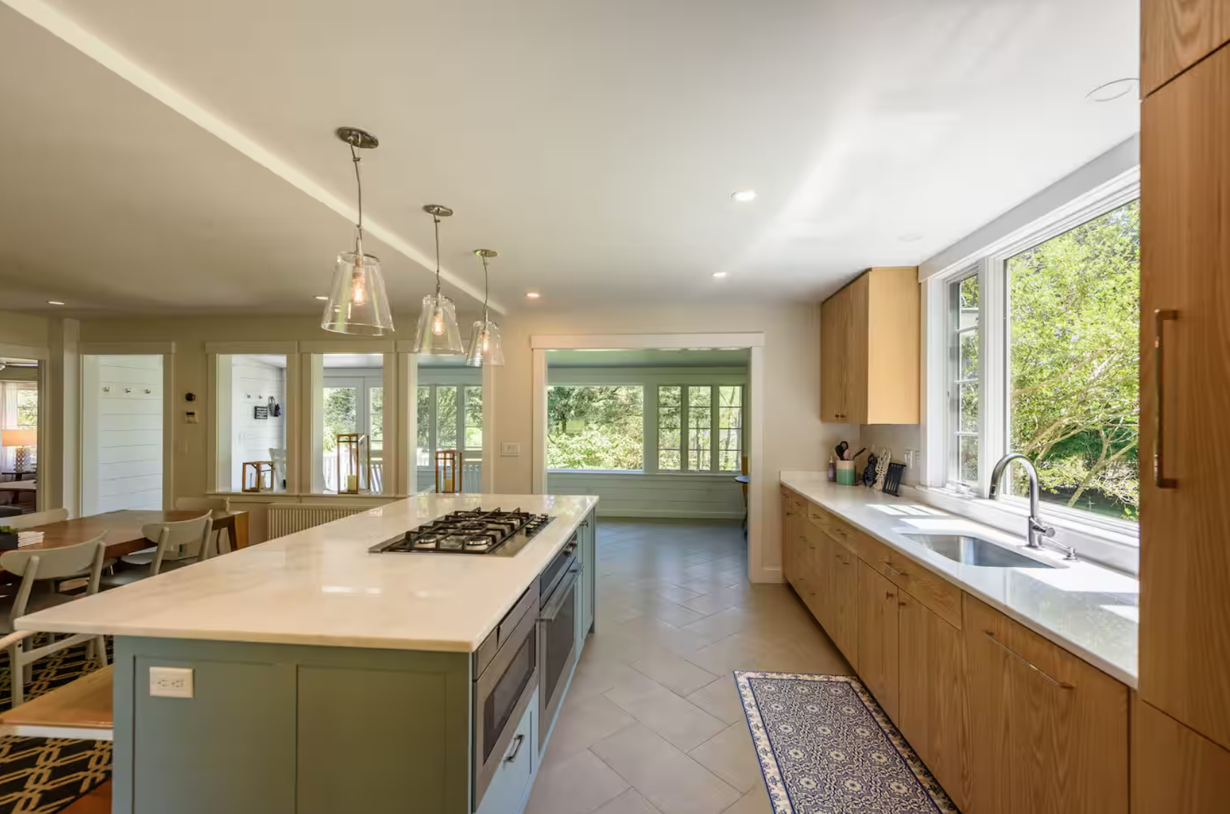 Bright kitchen with large window, wooden cabinets, white countertops, central island with stovetop, dining area with chairs, pendant lights, and views of green trees outside.