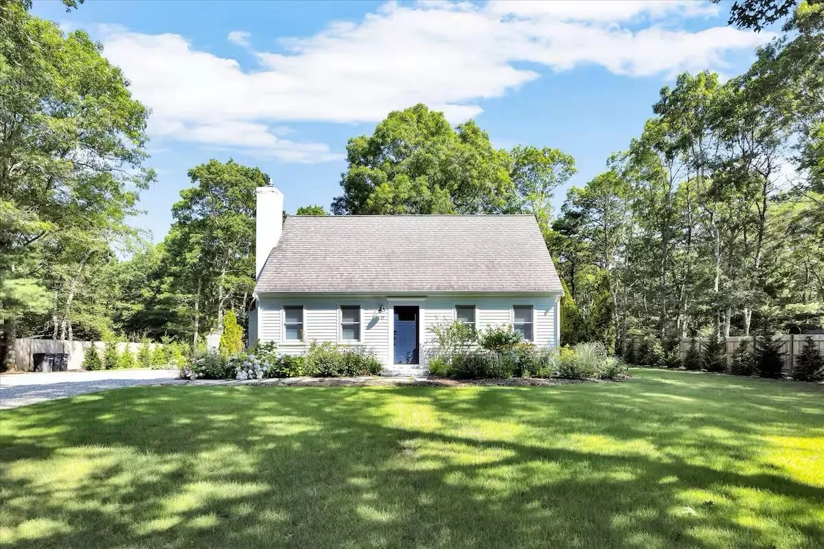 White house with gray roof surrounded by greenery and lawn under blue sky with clouds.