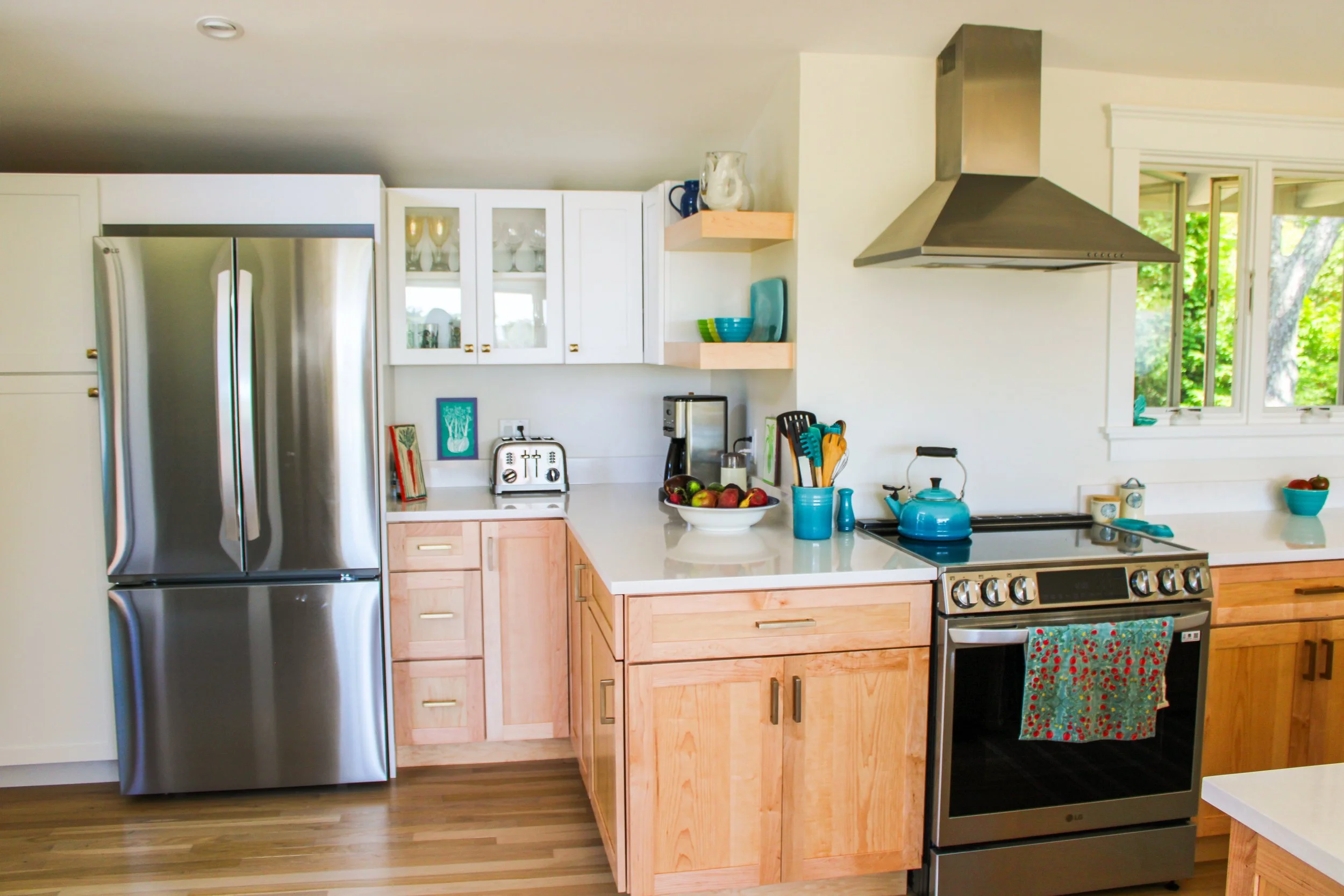 Kitchen with stainless steel refrigerator, light wood cabinets, white countertops, blue teapot on stove, and window showing green trees outside.