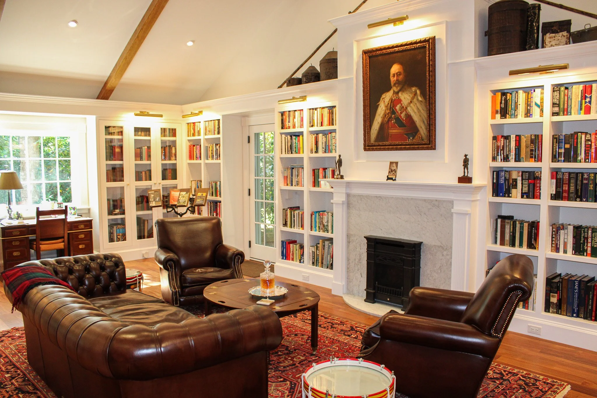 A cozy living room with a white fireplace, surrounded by built-in bookshelves filled with books. There are three leather armchairs and a leather sofa arranged around a round coffee table with a decanter and glasses. A Persian rug covers the wooden floor, and large windows let in natural light, showing green foliage outside. There is a portrait of a man in a regal uniform hanging above the fireplace.