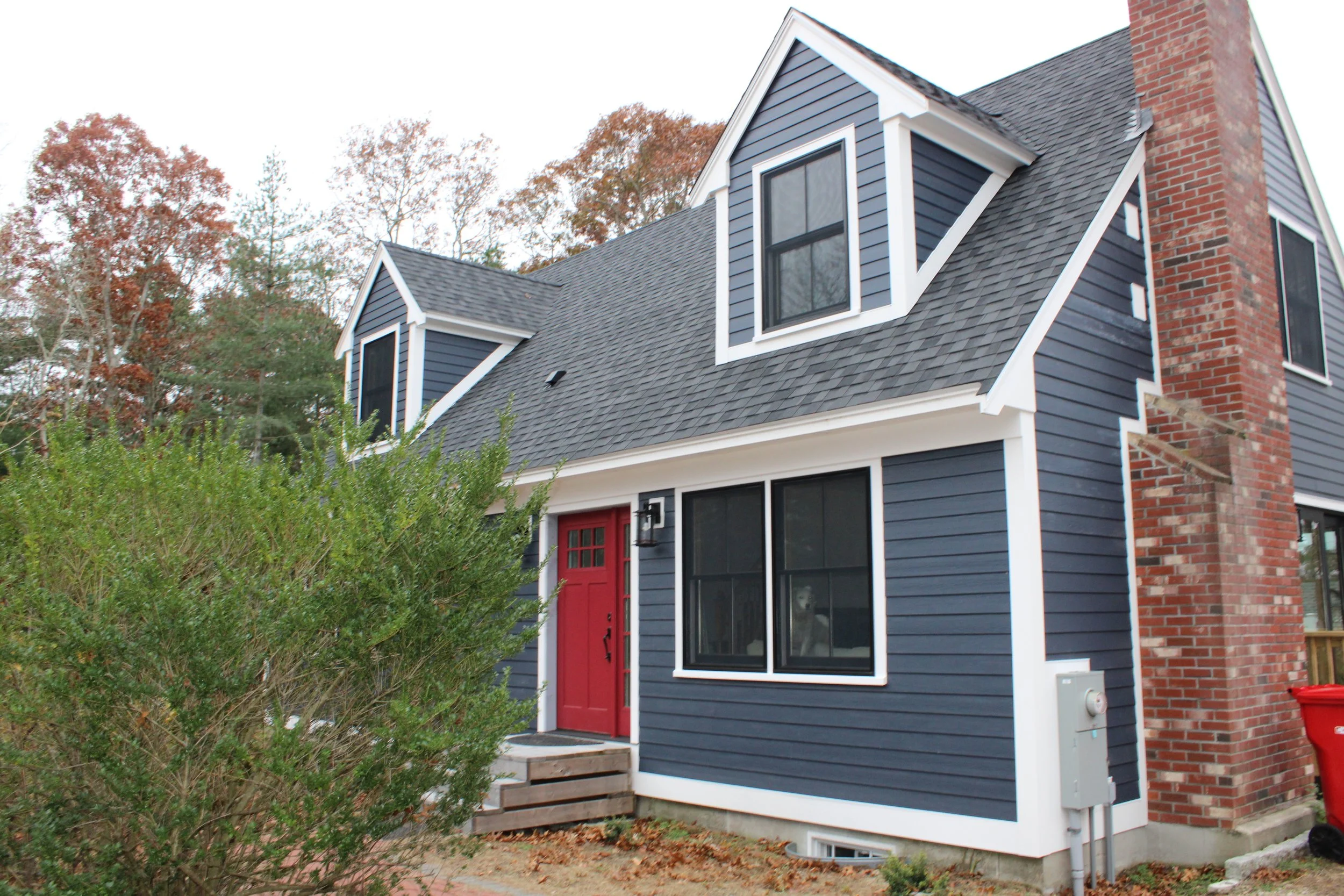 A two-story house with dark blue siding, white trim, and a red front door. The house features a gabled roof, double-hung windows, and a brick chimney. There are steps leading up to the front door and a garden with shrubbery in the front yard.
