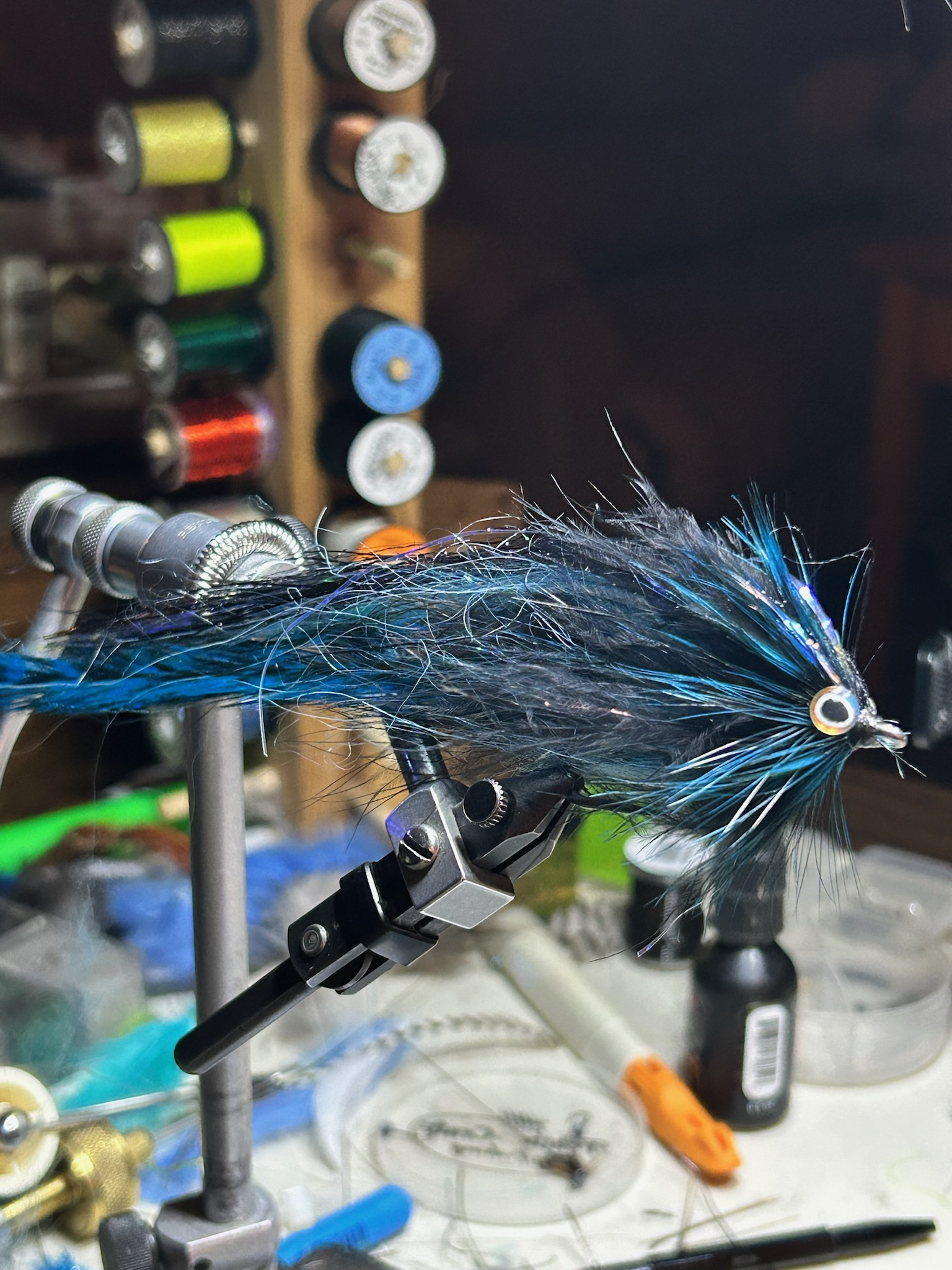 Close-up of an artificial fishing lure with blue and black feathers mounted on a vise, with a fishing hook. Background shows spools of colorful threads on a wooden rack.