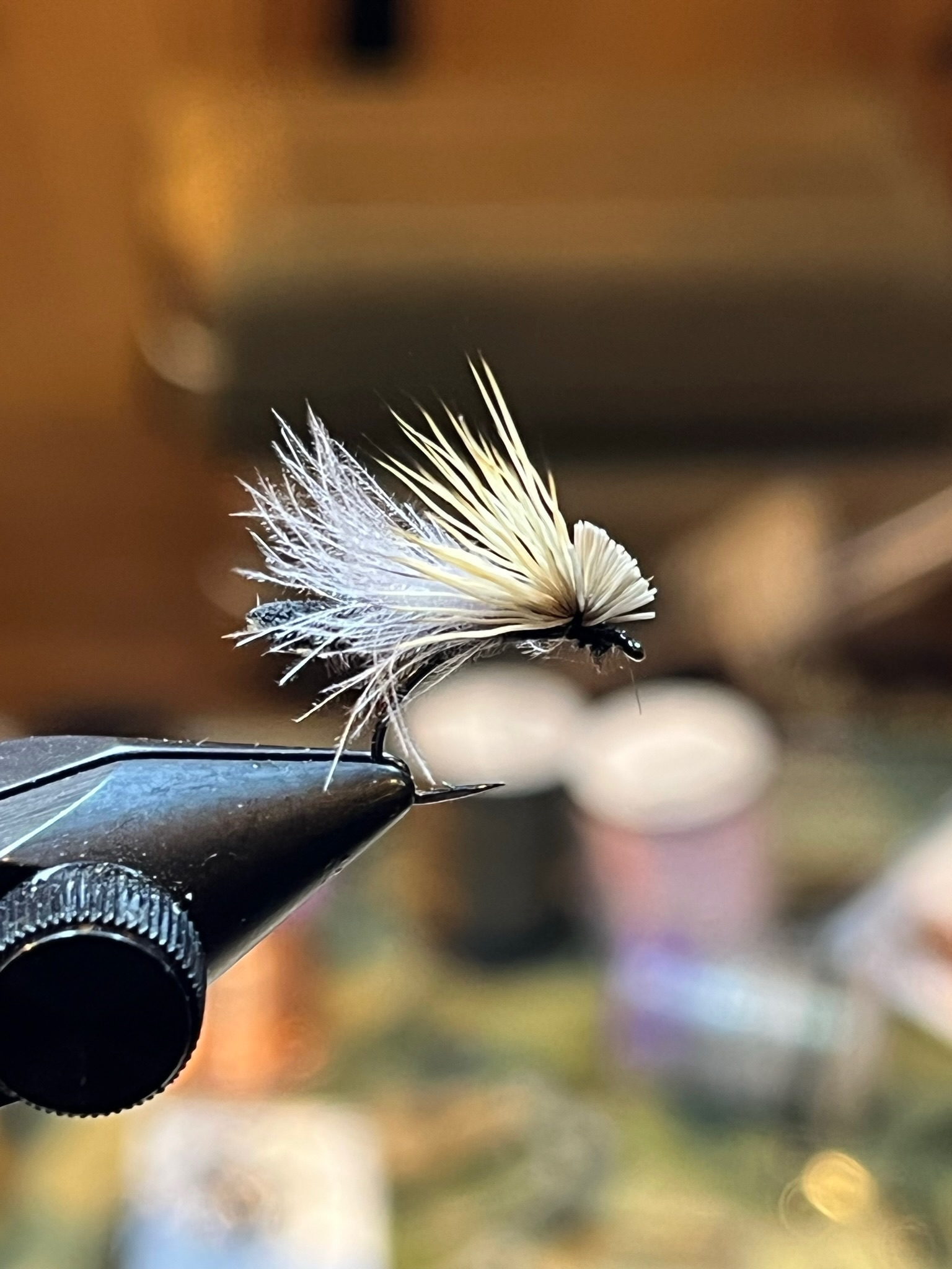 Close-up of a fishing hook with a white and black dry fly fishing lure, showing detailed feather and hair fibers, against a blurred background.