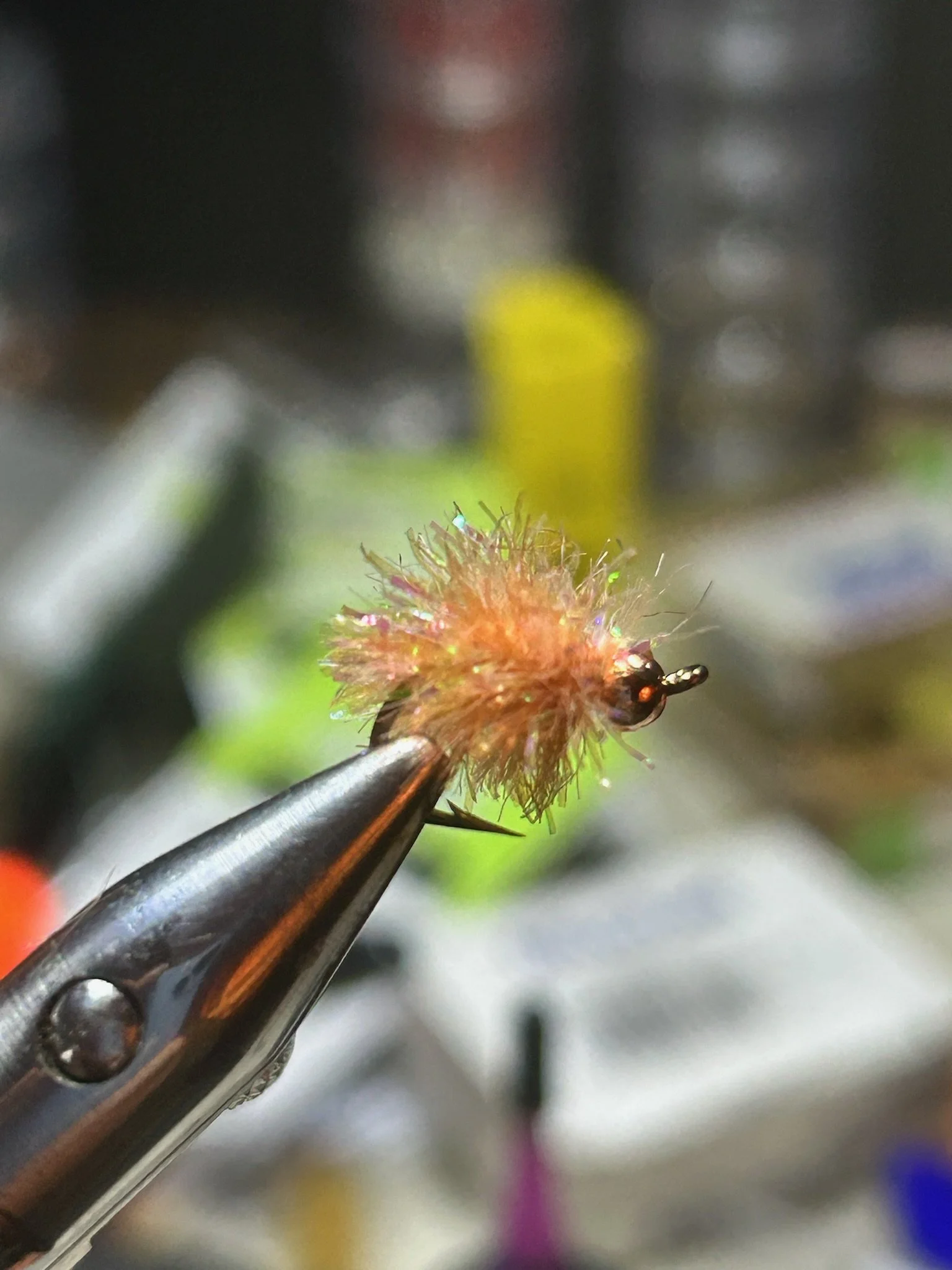 Close-up of a fly fishing lure with feathers and a hook, held by tweezers, with a blurred background of various colors.