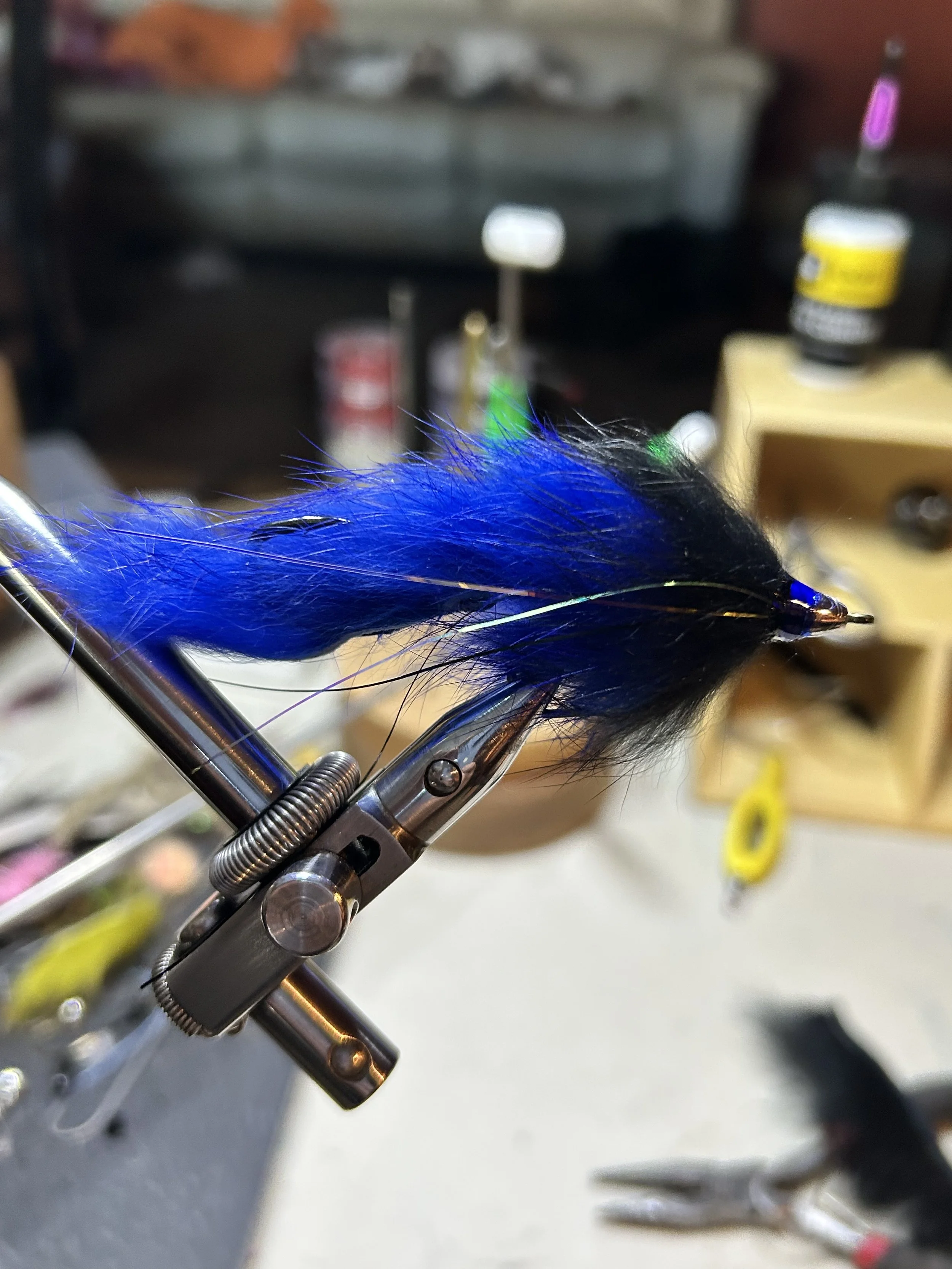 A close-up view of a fishing fly with blue and black feathers, held in a metal clamp in a workshop.