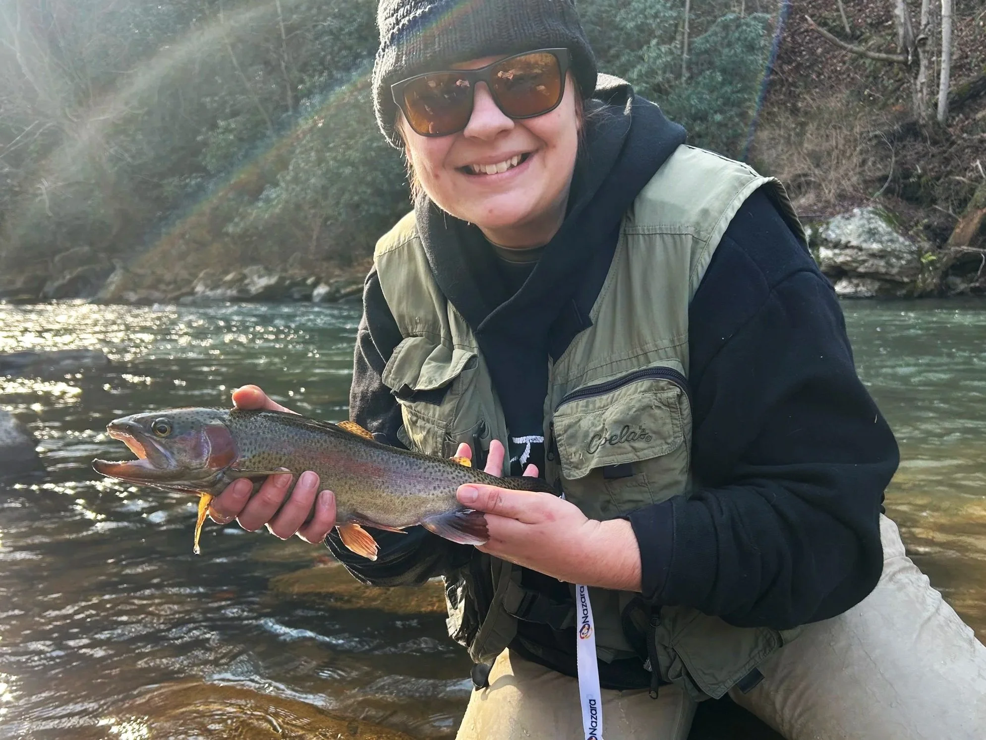 Person in outdoor gear holding a rainbow trout in a river, with trees and rocks in the background, and sunlight reflecting on the water.