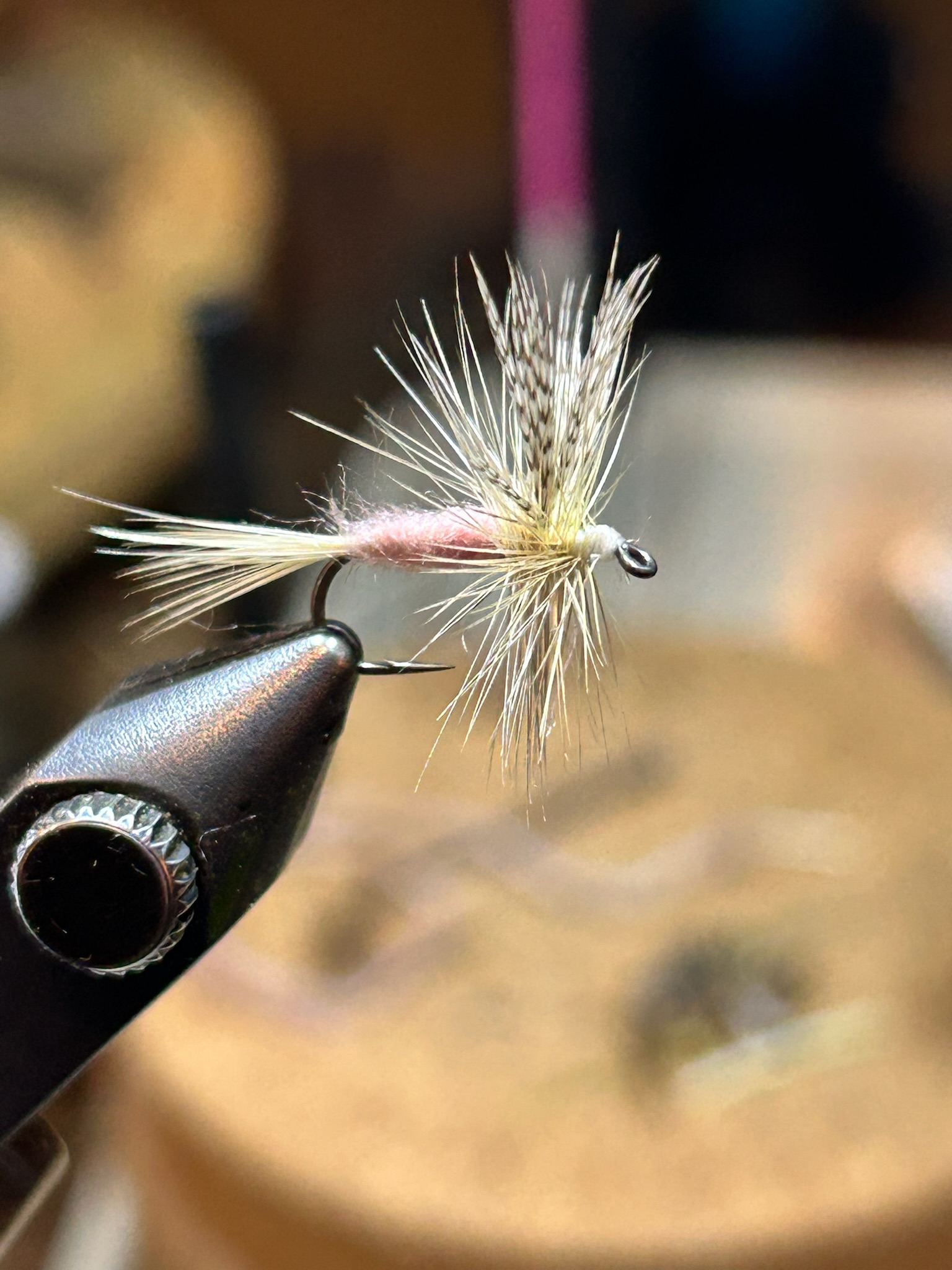 Close-up of a fishing fly with feathers and fur, held by a black vise or clamp, set against a blurred background.