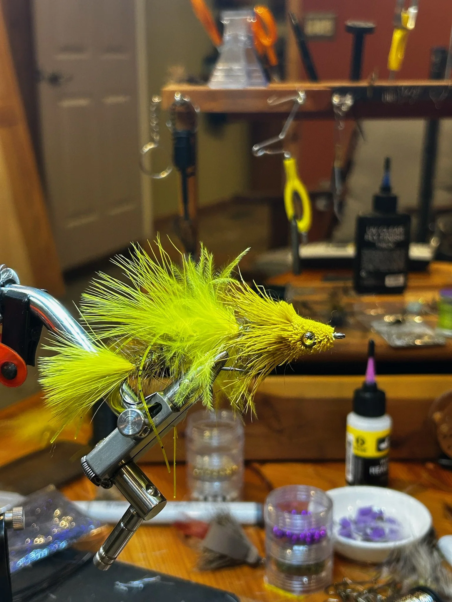 A bright yellow and green fishing fly being tied on a vise in a fly-tying workspace with various tools and materials in the background.