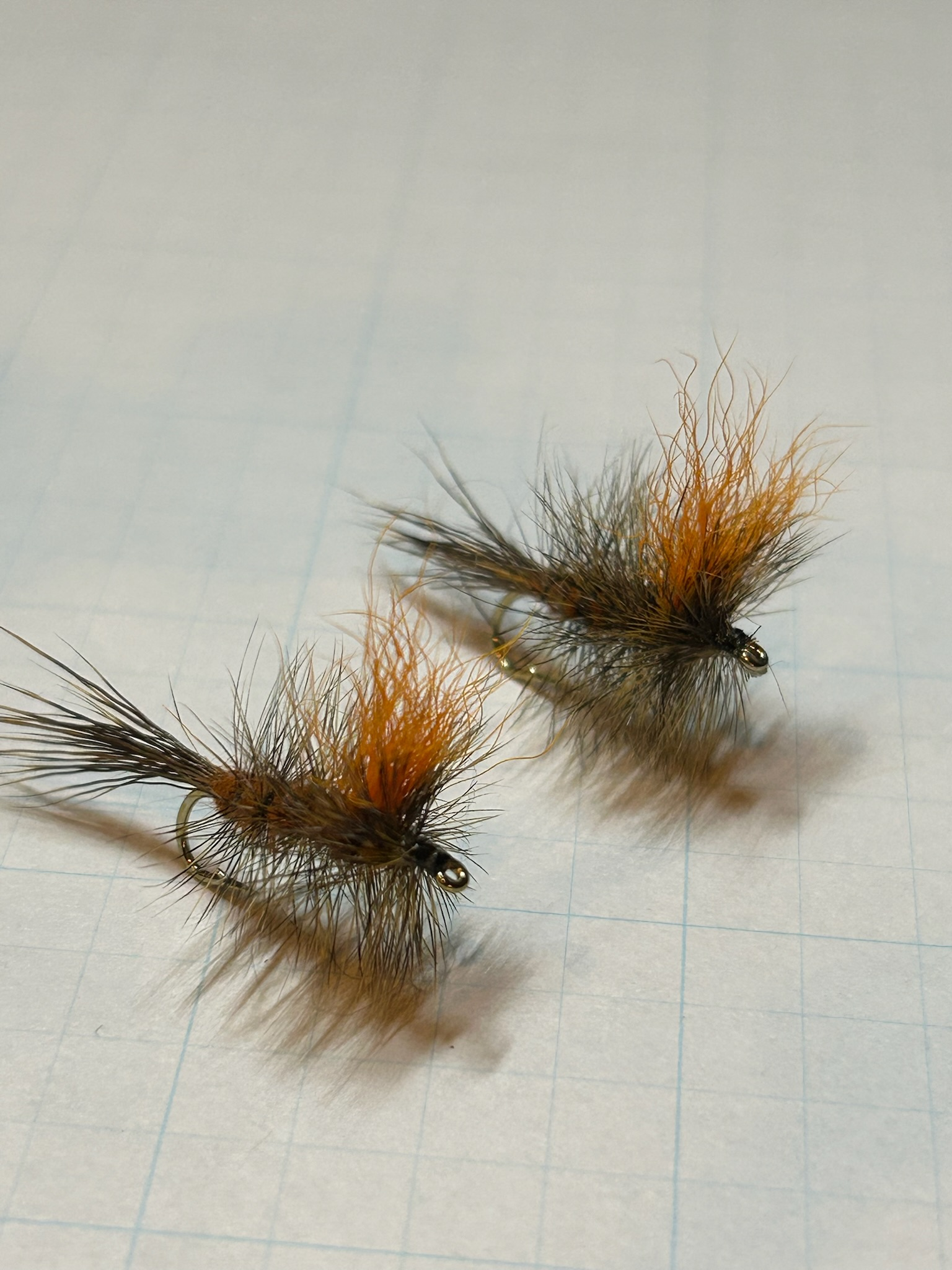 Two fishing flies with orange and brown feathers on a white paper with blue grid lines.