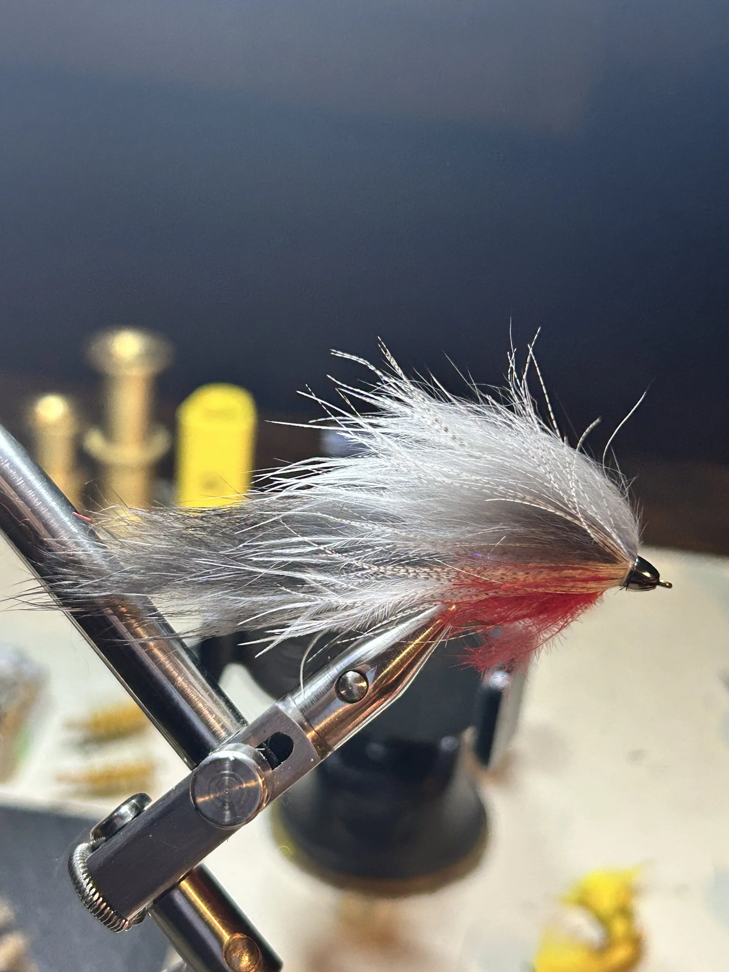 A fishing fly with white and red feathers mounted on a metallic clamp, with a blurred background including some yellow and silver objects.