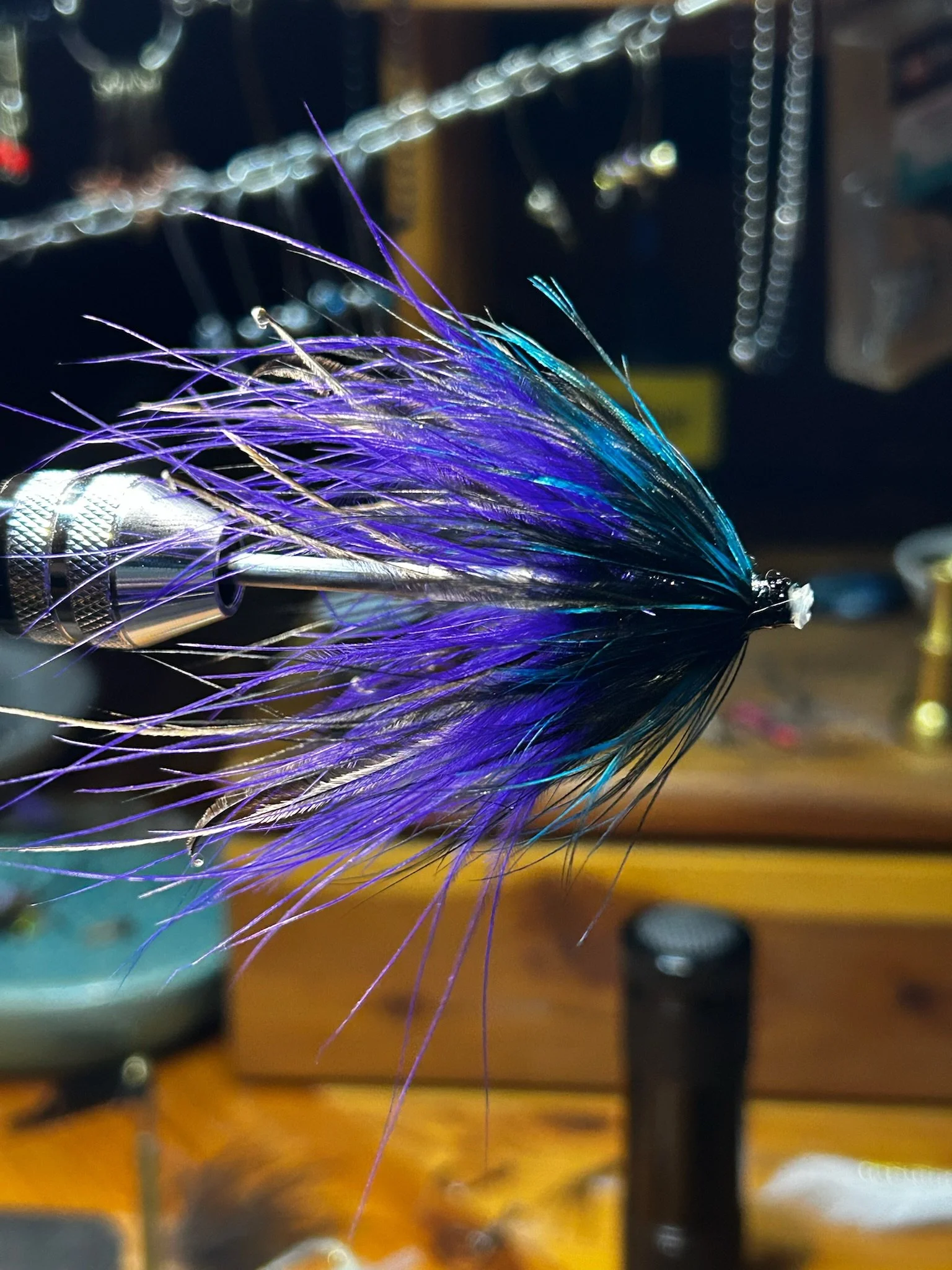 A close-up of a fishing fly with vibrant purple, blue, and black feathers, mounted on a metal hook, and held in place with a vice grip, in a fly-tying workspace.