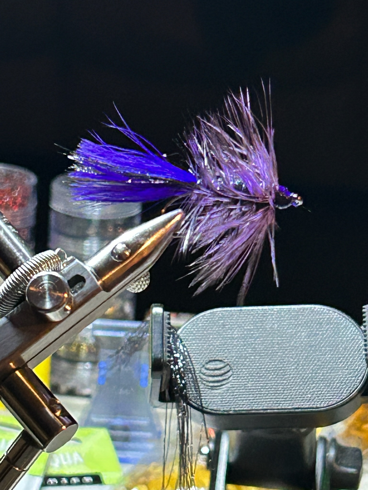 Close-up of a purple and blue fishing fly held in a pair of metallic pliers, with a vise and small containers in the background.