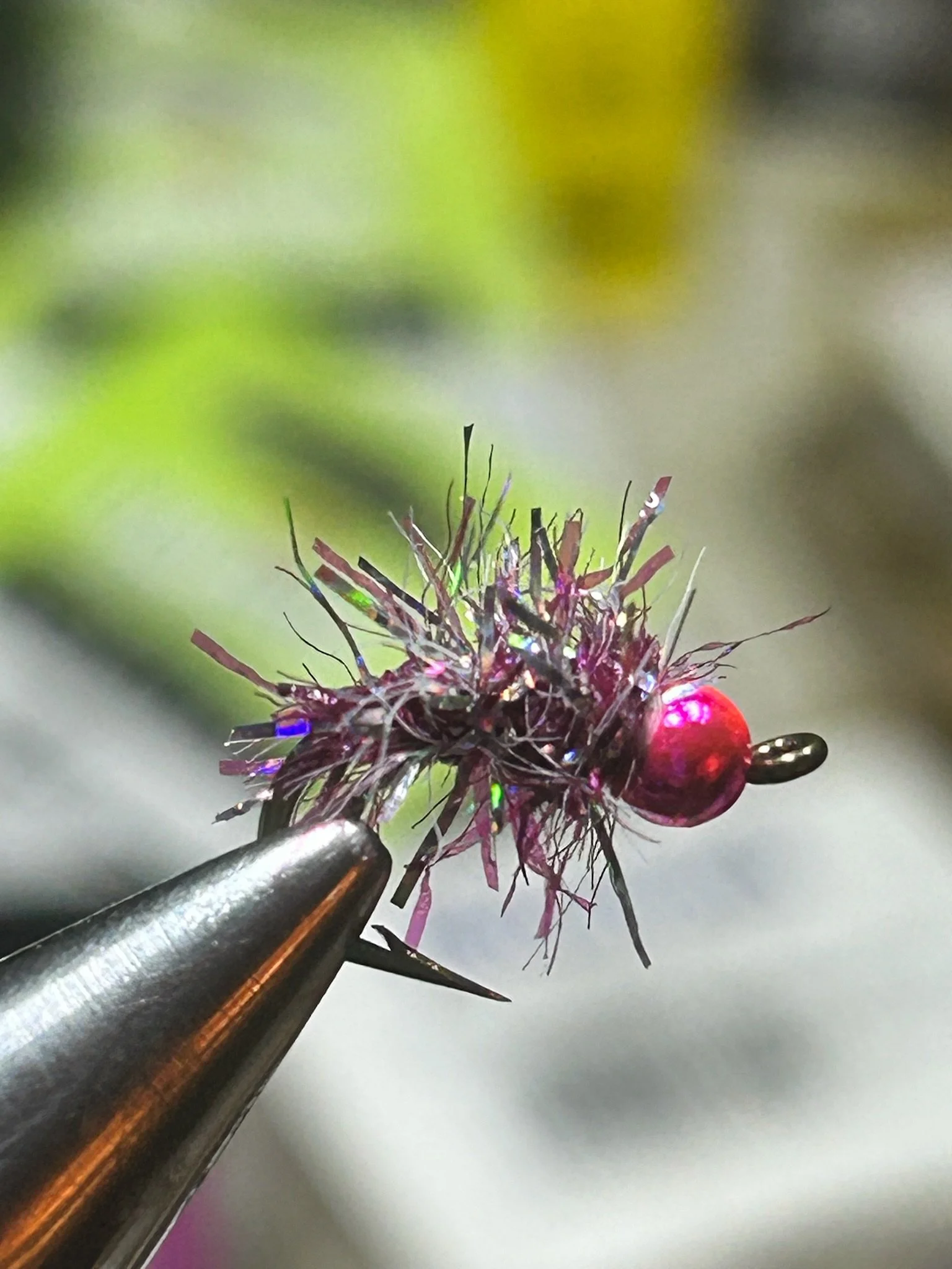 Close-up of a small, colorful fishing lure with black, purple, and iridescent strands, and a shiny red bead, held by a metallic tool with a blurred green and gray background.