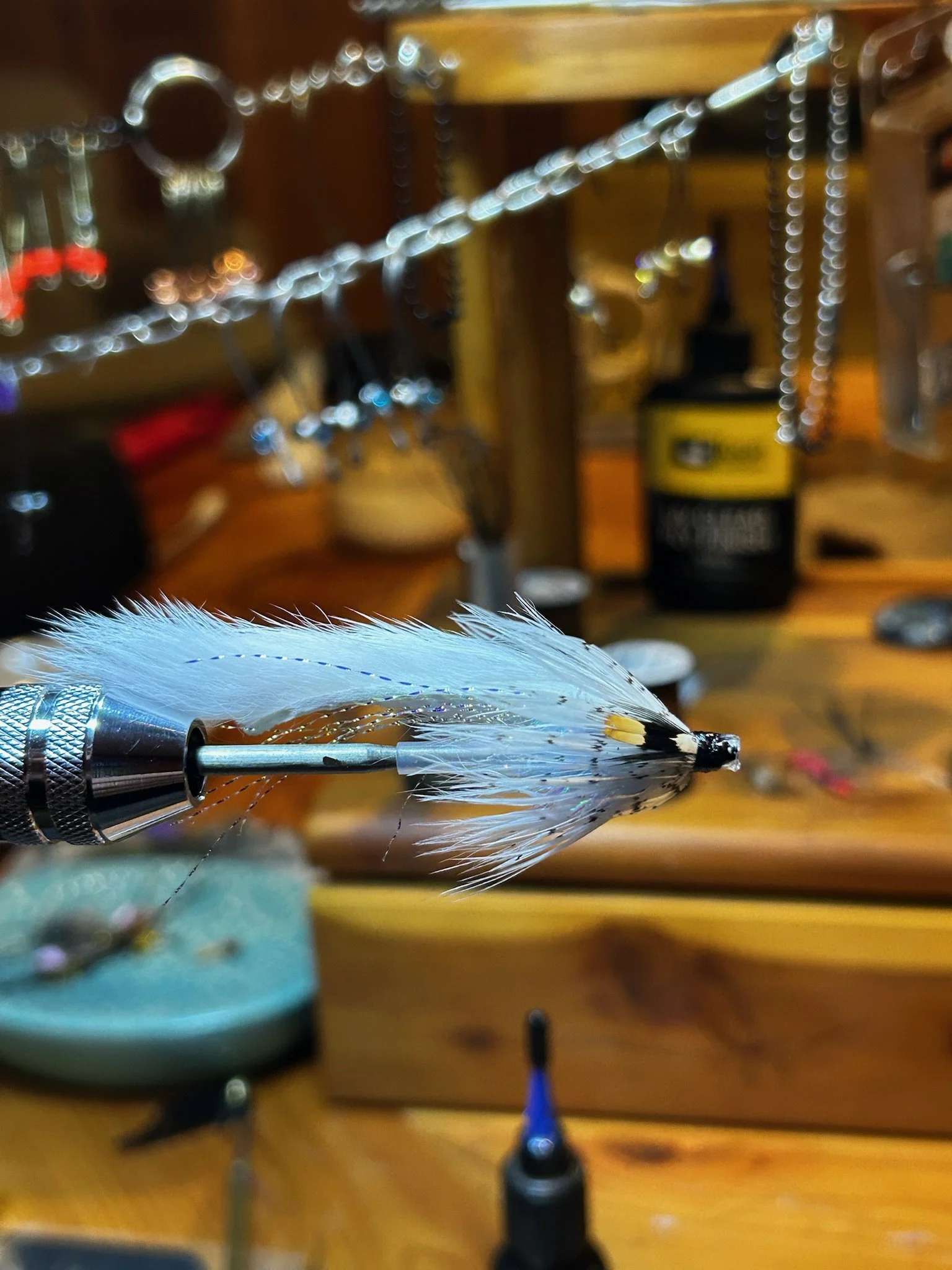 Close-up of a fishing lure with white feathers attached, held in a metal holder on a wooden workbench with agility tools and supplies in the background.