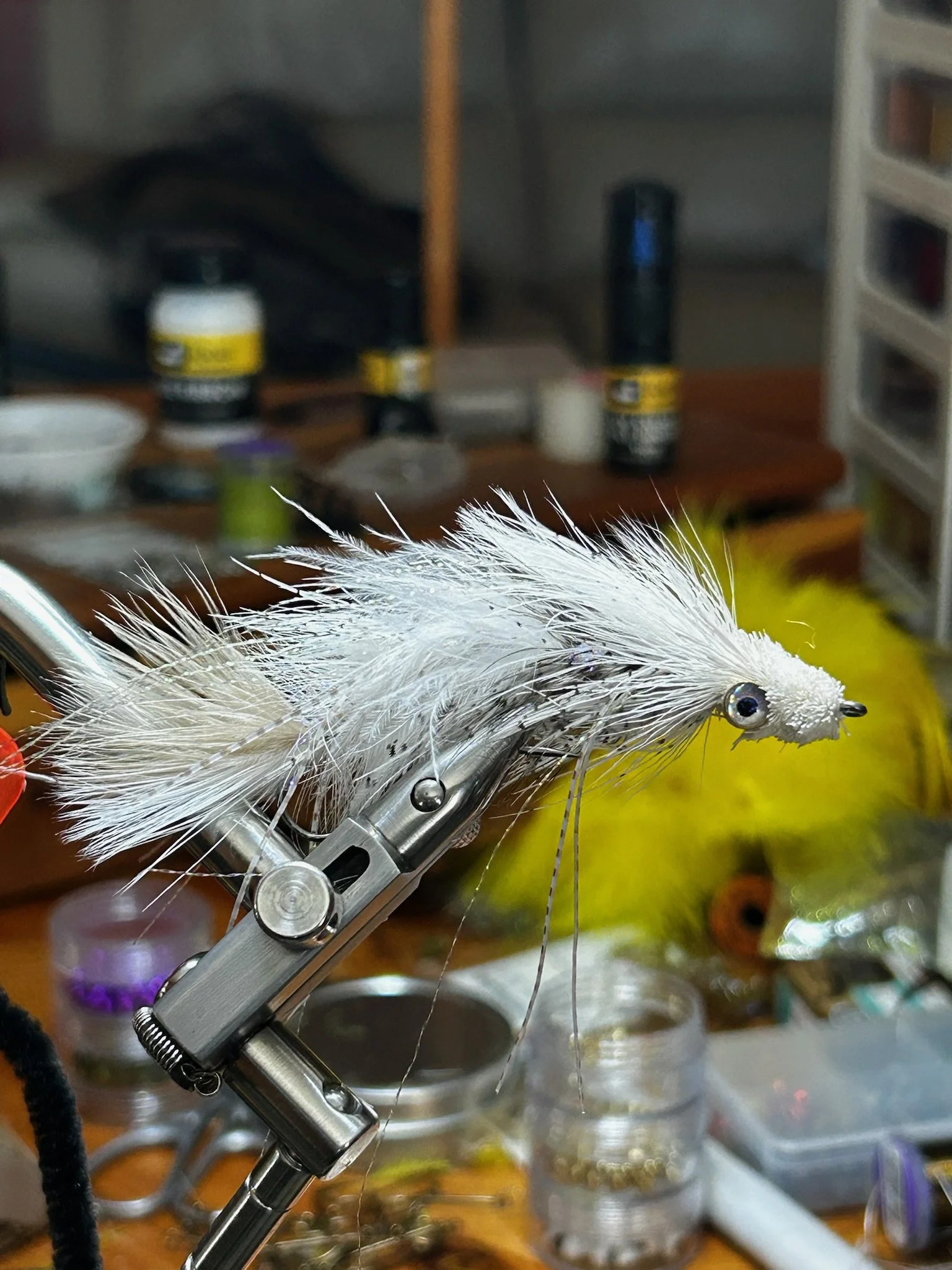 Close-up of a detailed fishing lure or fly with white and gray feathers held by a metal clamp on a workbench surrounded by sorting containers and tools.