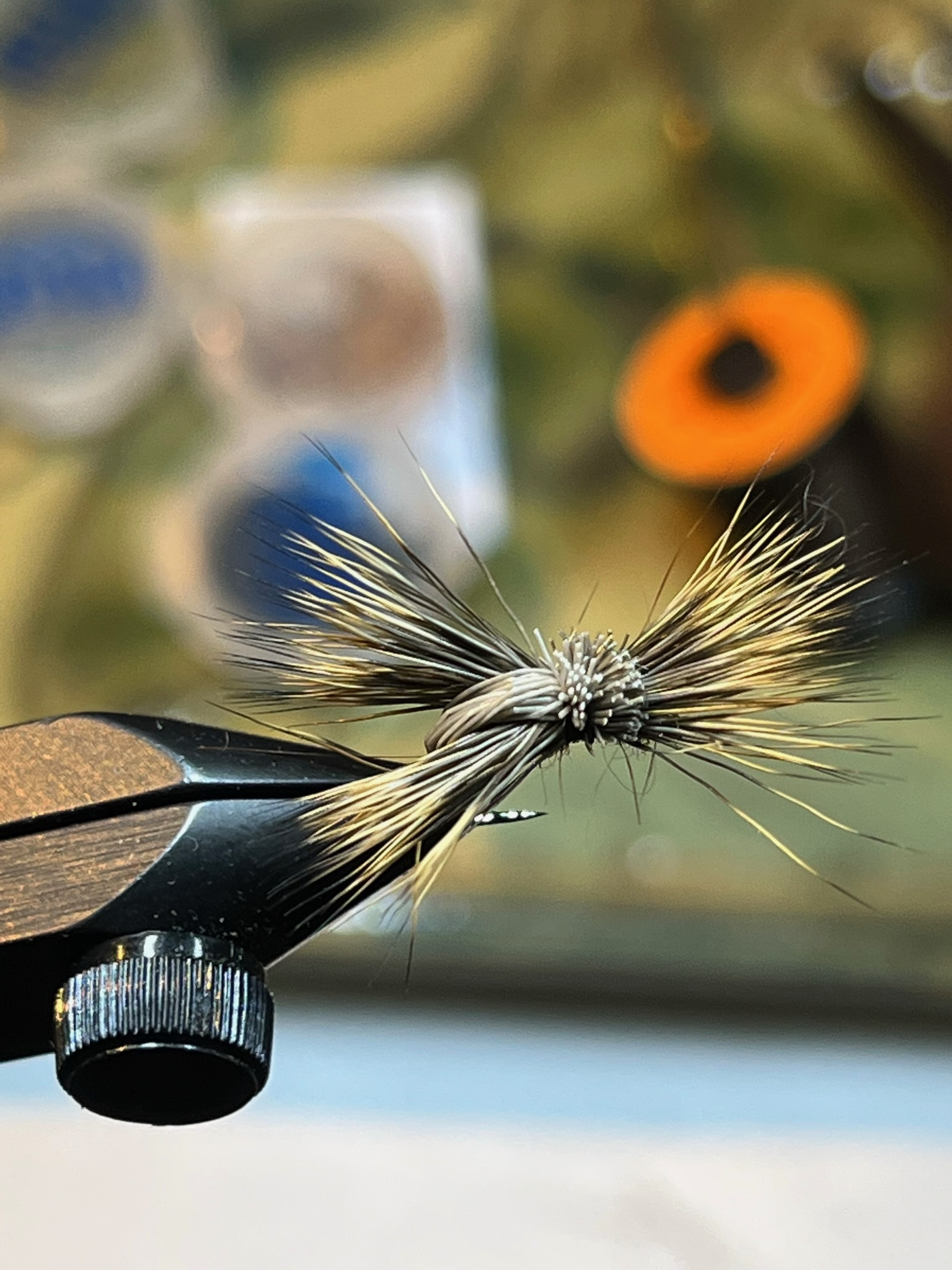Close-up of a fly-fishing artificial fly lure held in a vise, with a blurred background of outdoor items and a sunflower.