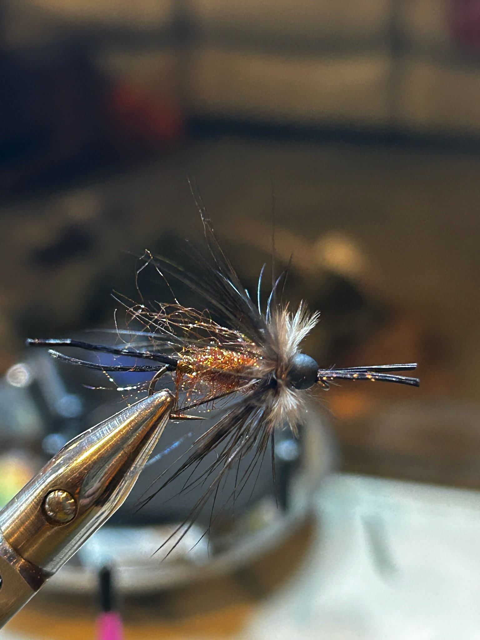 Close-up of a fishing fly with black, brown, and glittering gold elements, held by a metallic vise.
