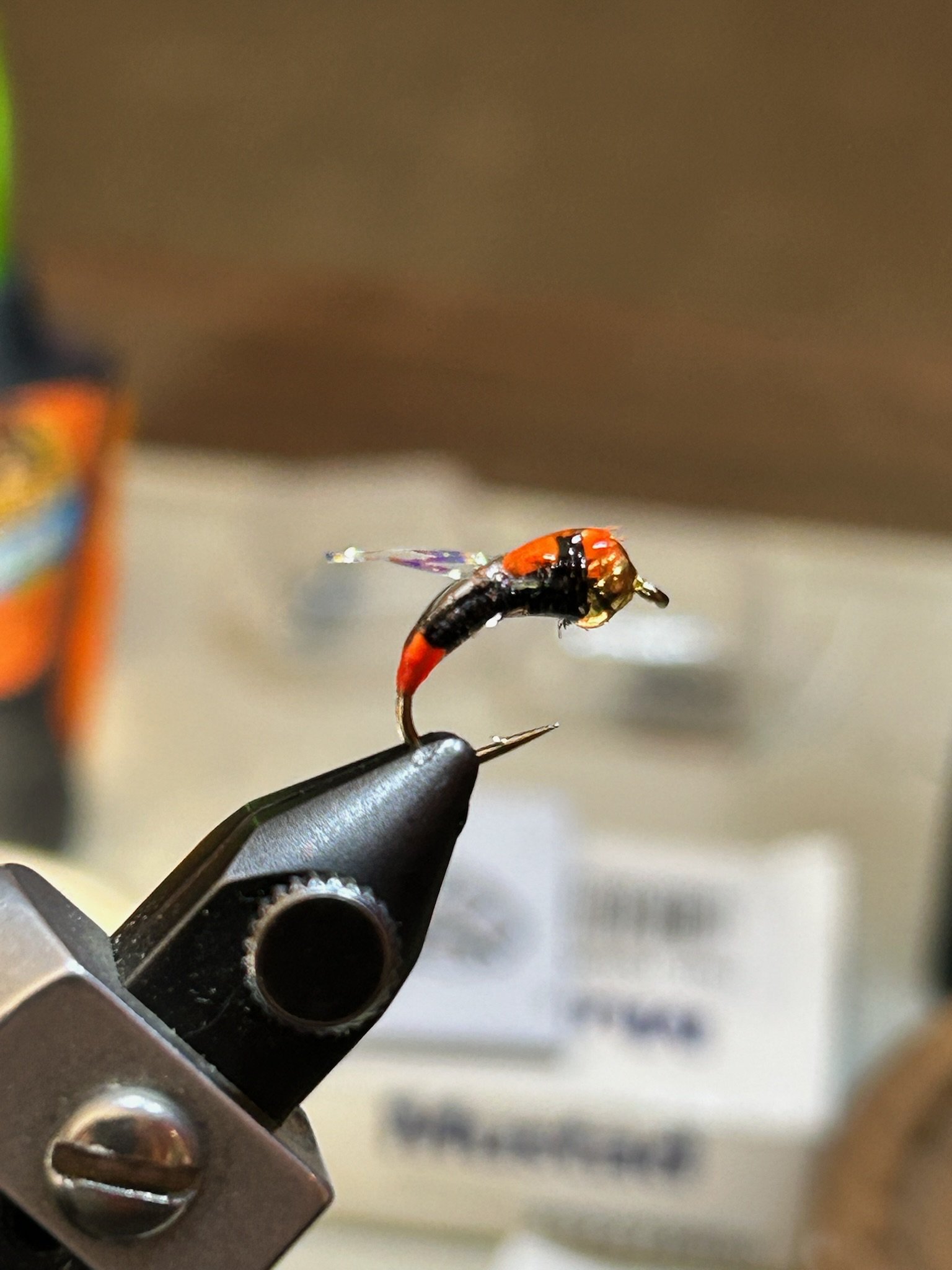 Close-up of a fly fishing lure with a red and black hook, held in a vise over a blurred background.
