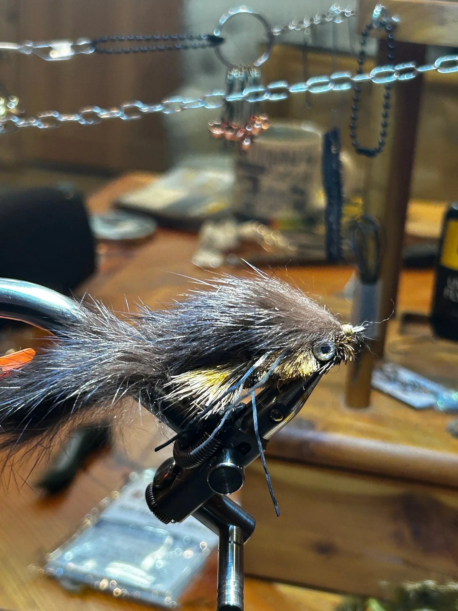 A fishing fly with black and yellow feathers held in place by a black clamp, with a blurred background of a craft workspace.